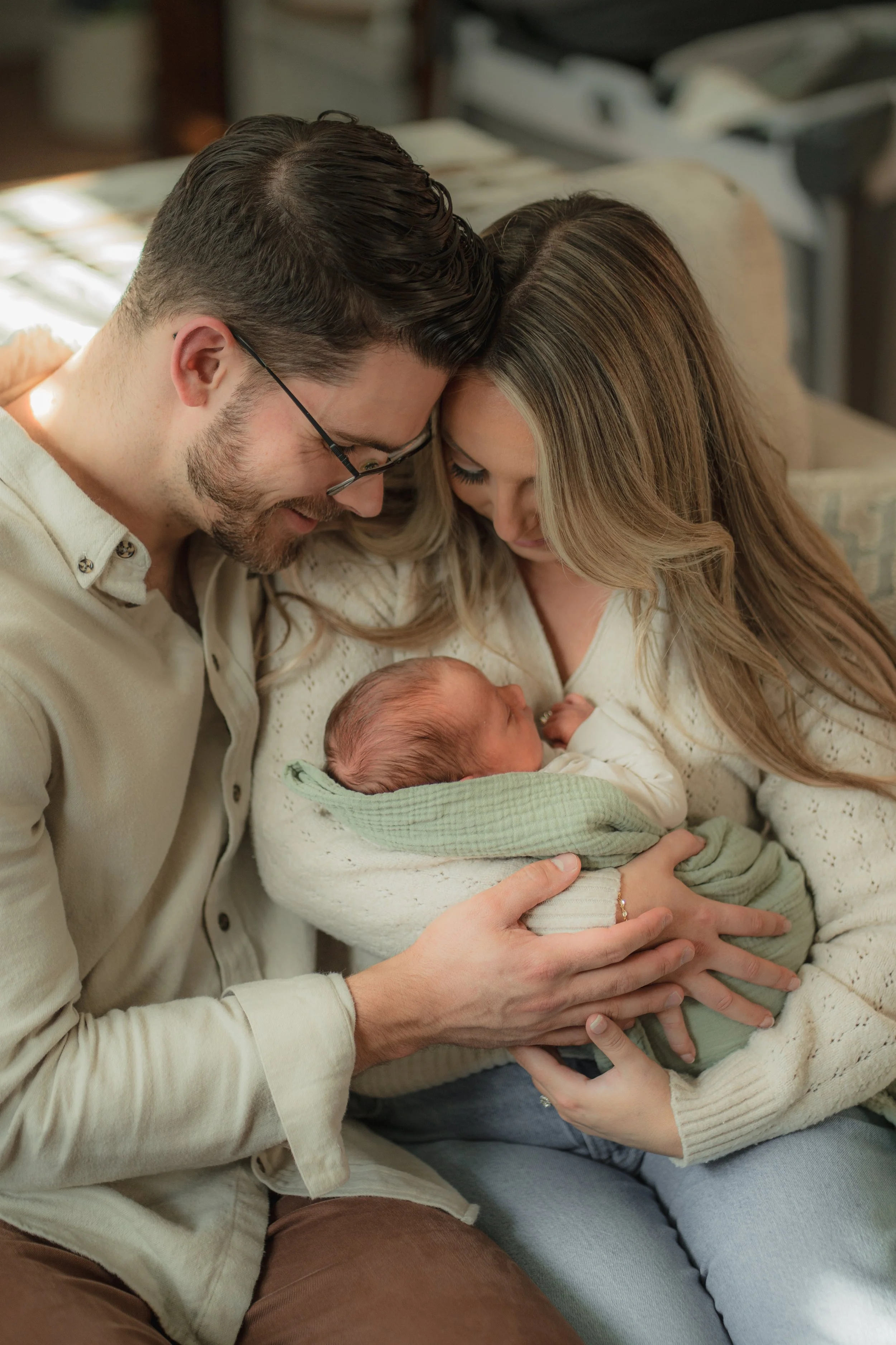 A family of three, including a newborn baby, sitting close together and looking at each other lovingly in a cozy setting.