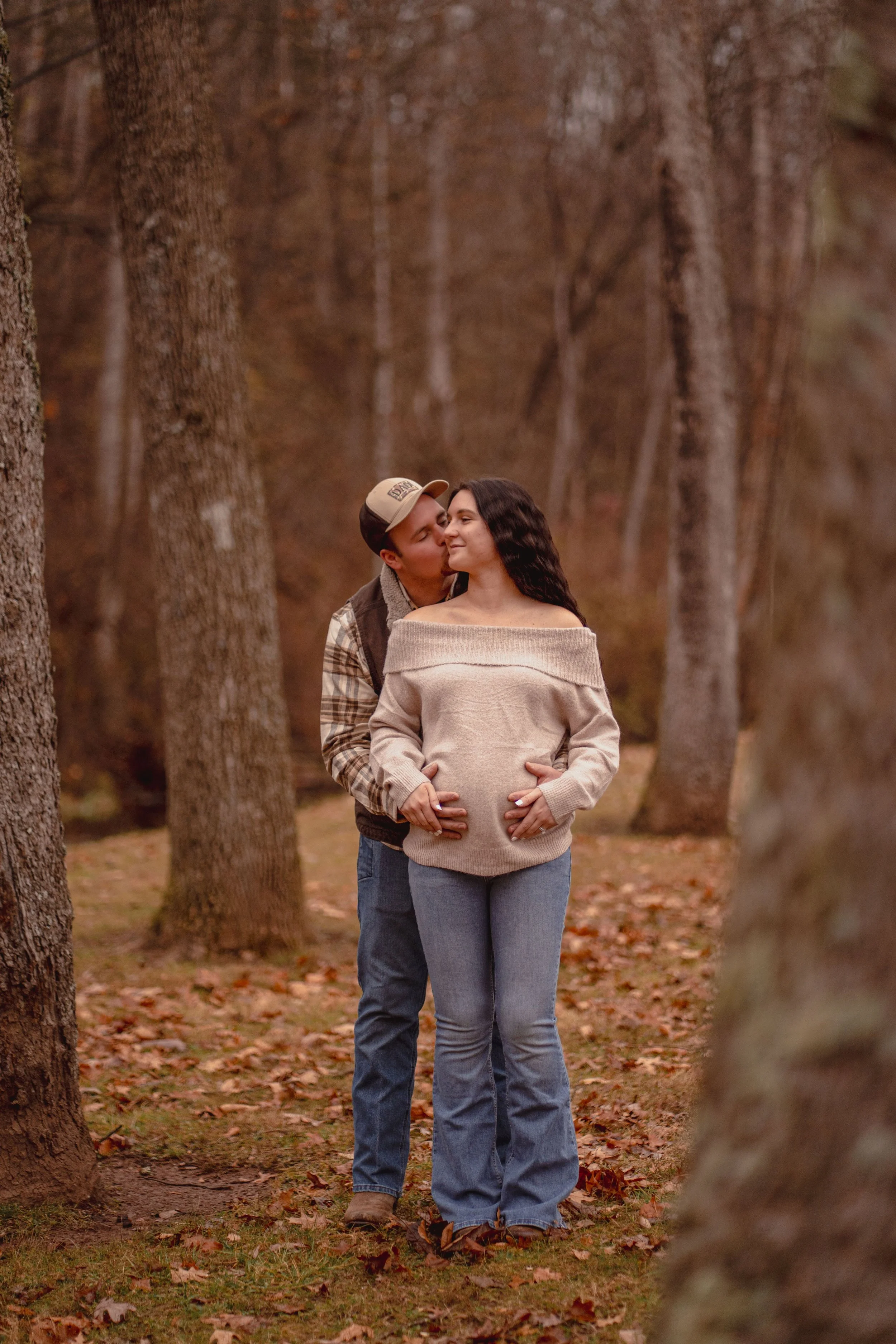 A couple, with the man kissing the woman's cheek, stands in an autumn forest, the woman being visibly pregnant.