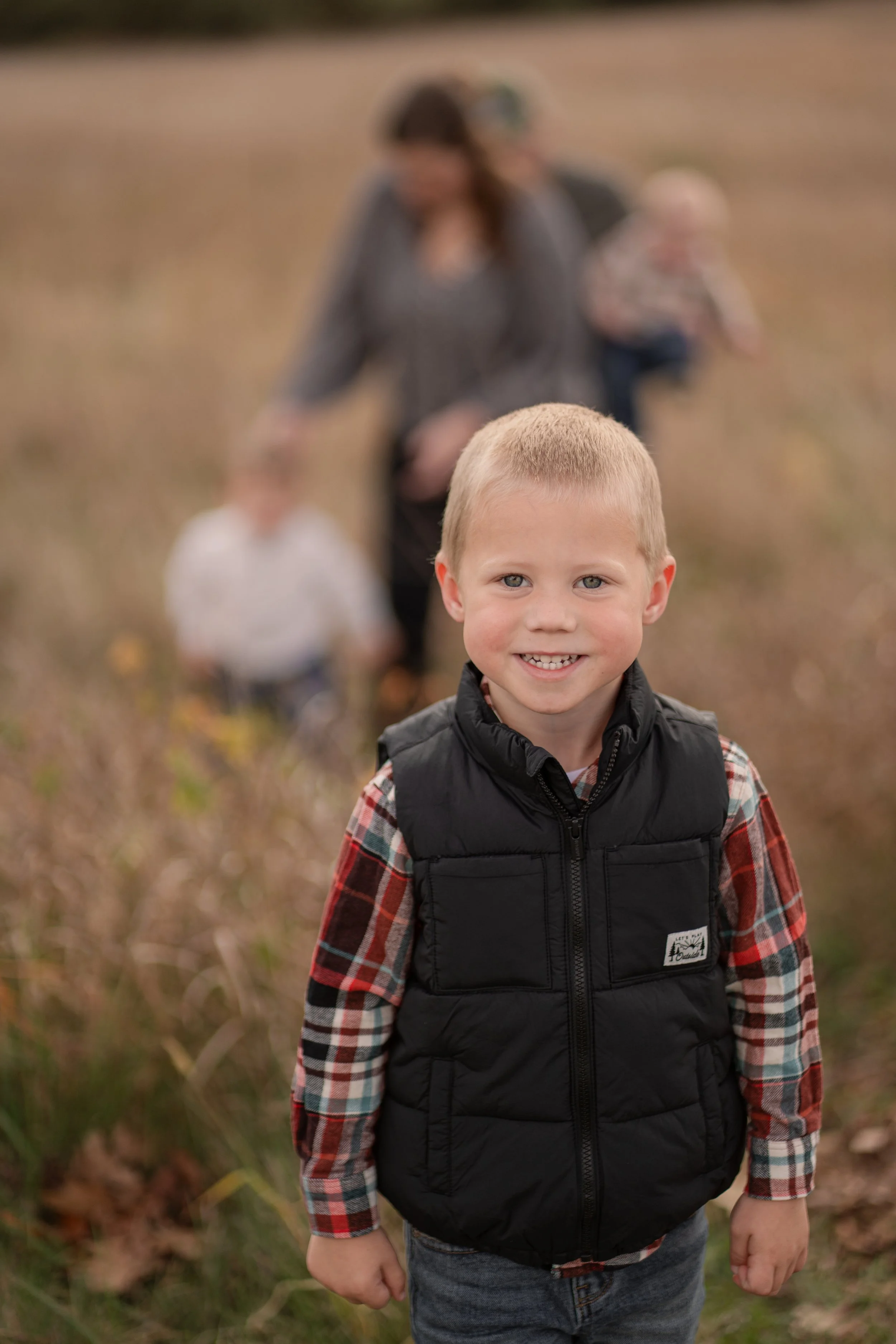 Young boy smiling in a field with family members blurred in the background.