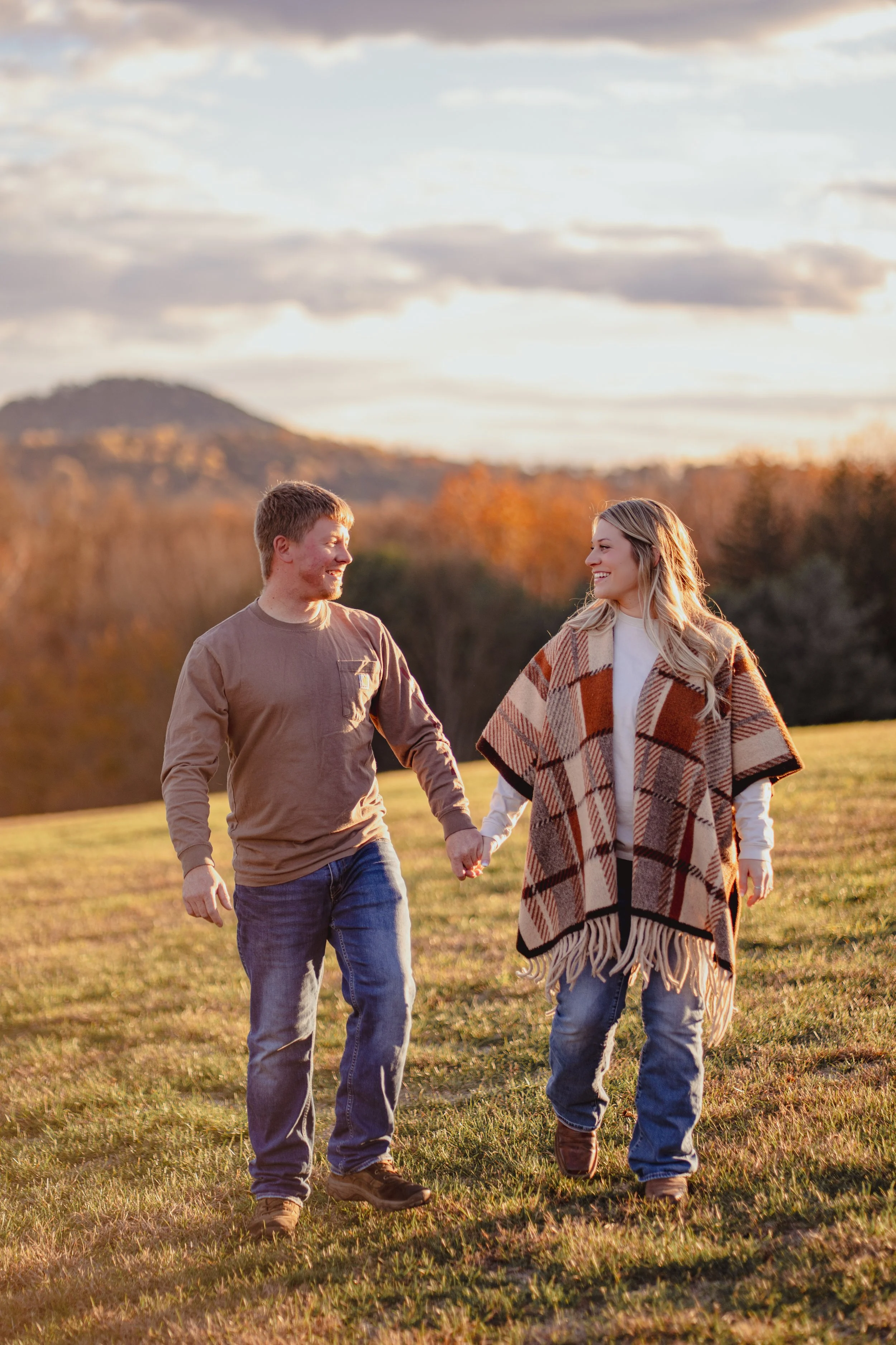 A couple walking hand in hand in a grassy field during sunset, with mountains and trees in the background.