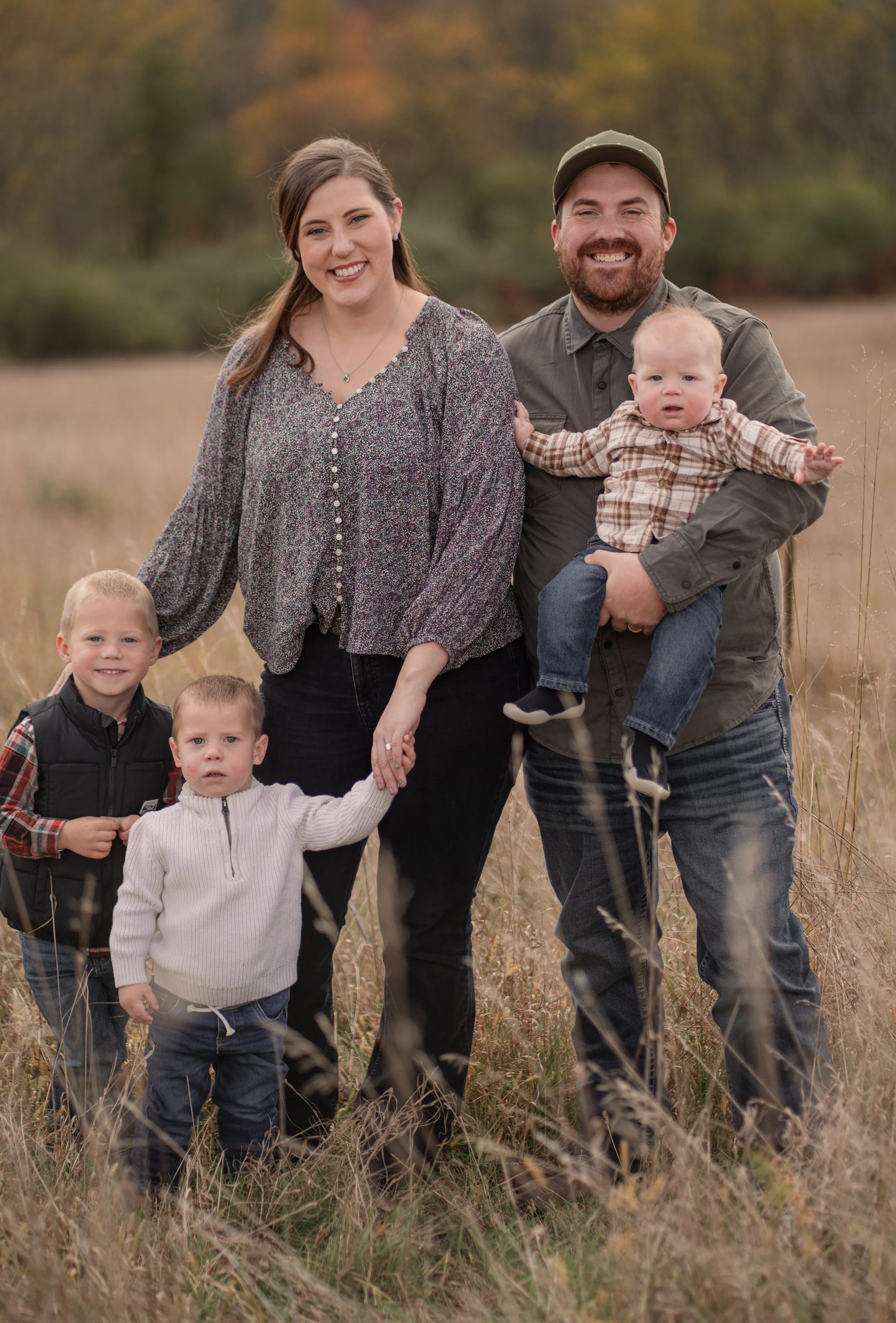 A family of six standing in a grassy field during autumn. The woman and man are smiling, with the man holding a baby boy. The woman is holding hands with two young boys, and another young boy is standing beside them.