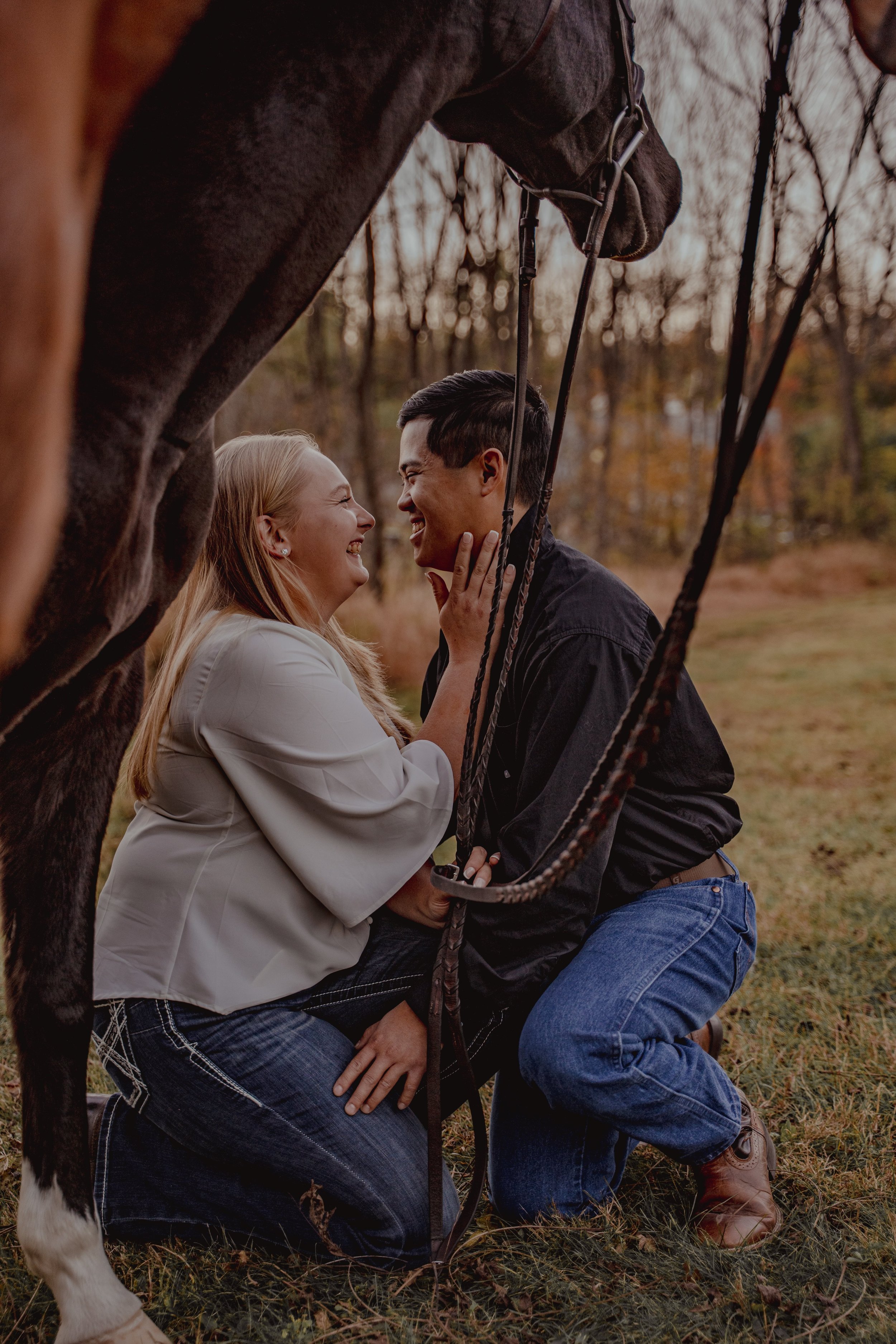 A couple kneeling close together, sharing a joyful moment under a horse's head, with fall trees in the background.