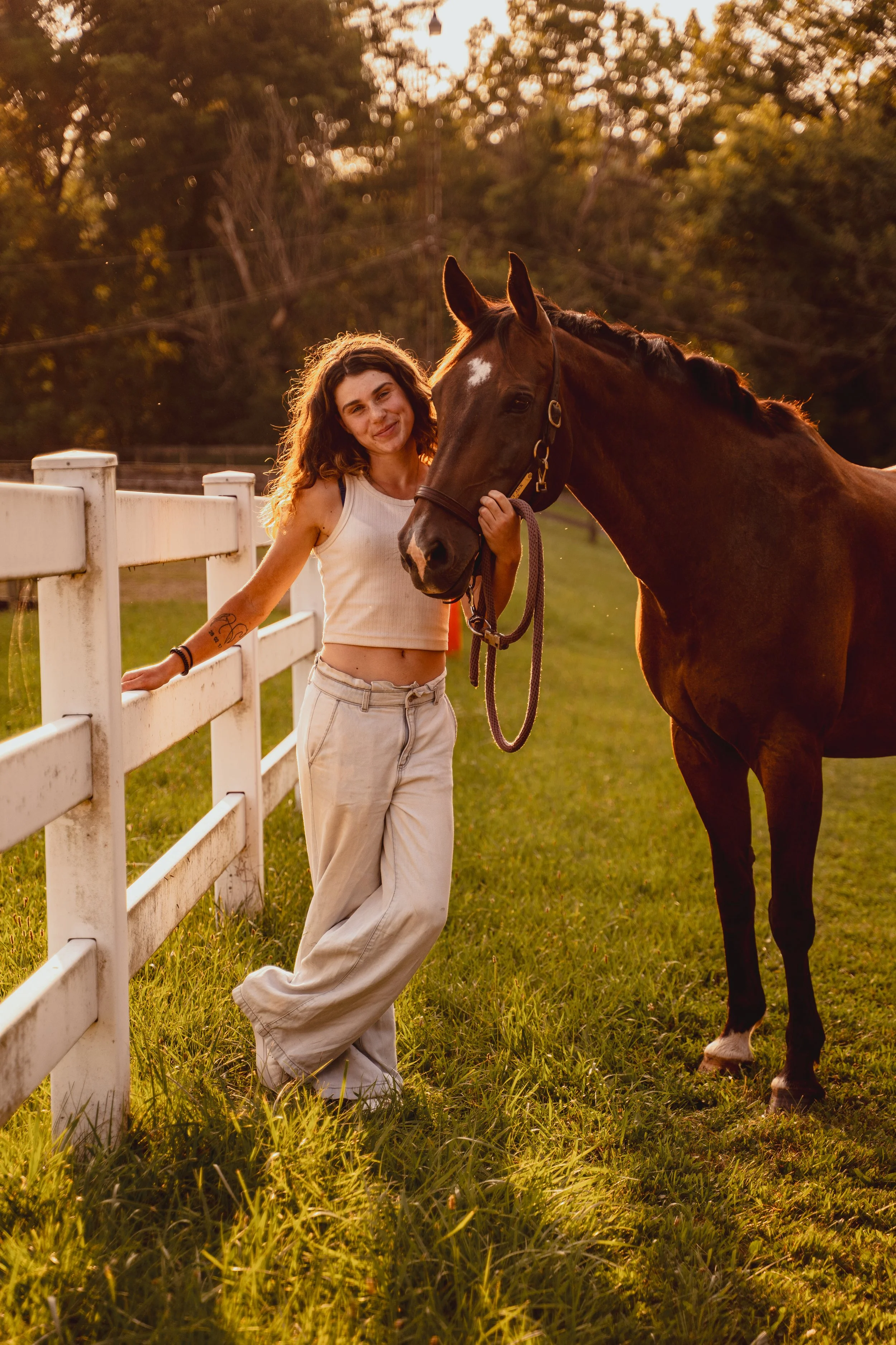 A young woman with curly brown hair, wearing a white sleeveless top and beige pants, standing next to a brown horse with a white stripe on its face, holding the horse's bridle in a grassy field with a white fence and trees in the background during su