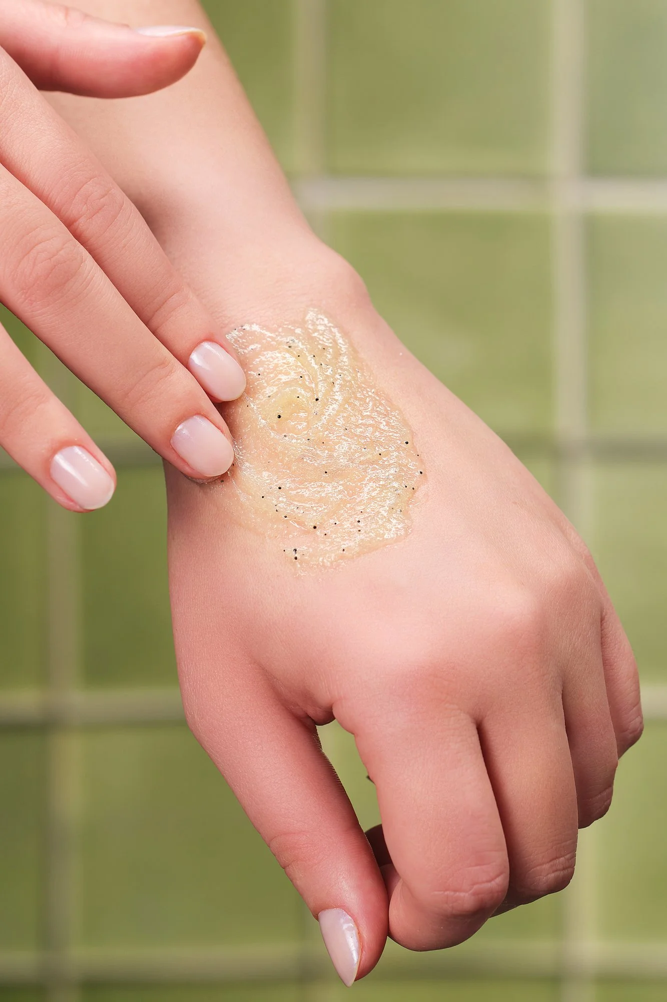 Pessoa aplicando um produto esfoliante na mão com as pontas dos dedos, fundo de azulejos verdes.