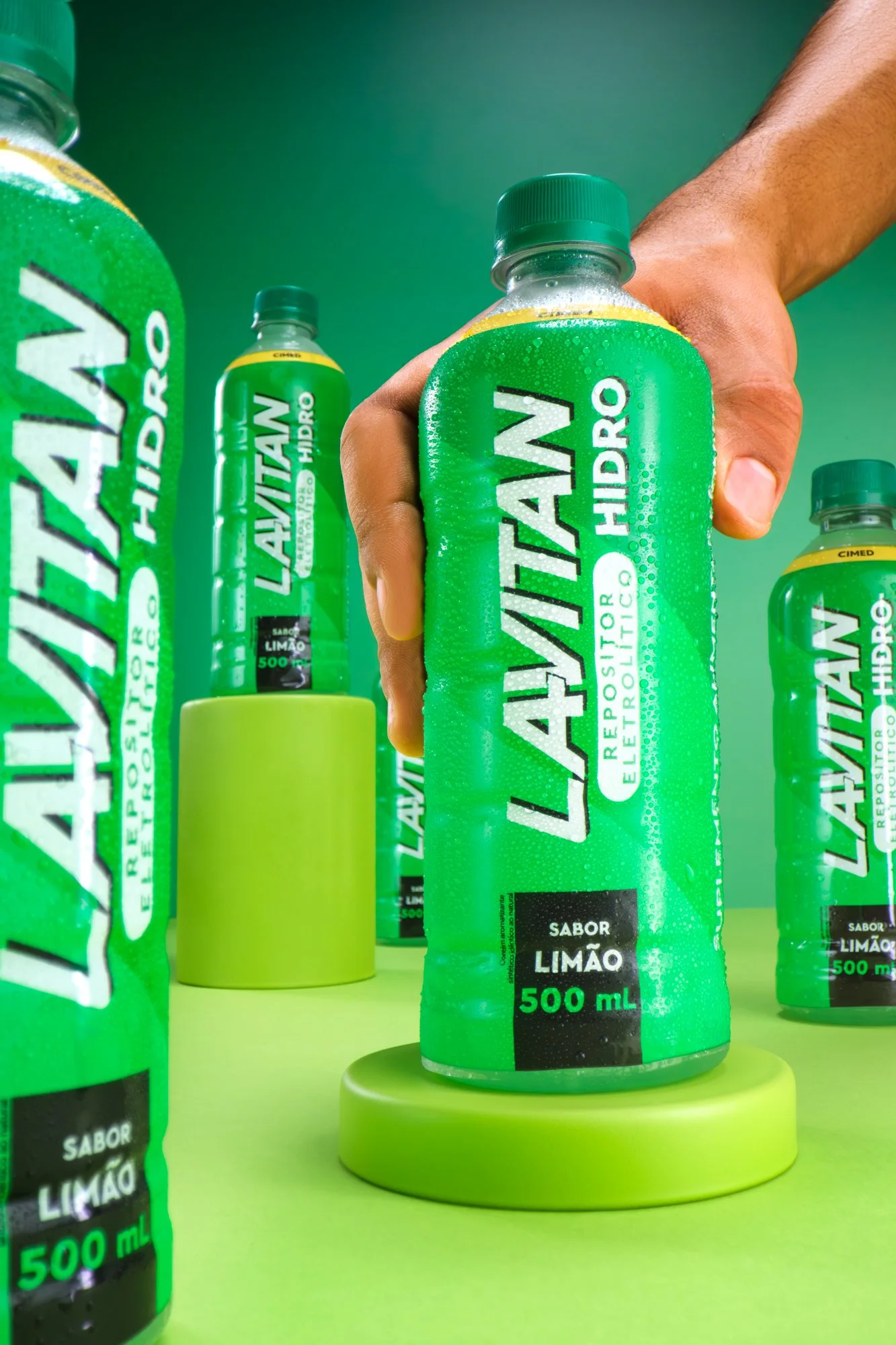 Imagem de várias garrafas verdes de refrigerante de limão, com uma pessoa segurando uma delas, ao fundo uma mesa verde e uma parede verde.