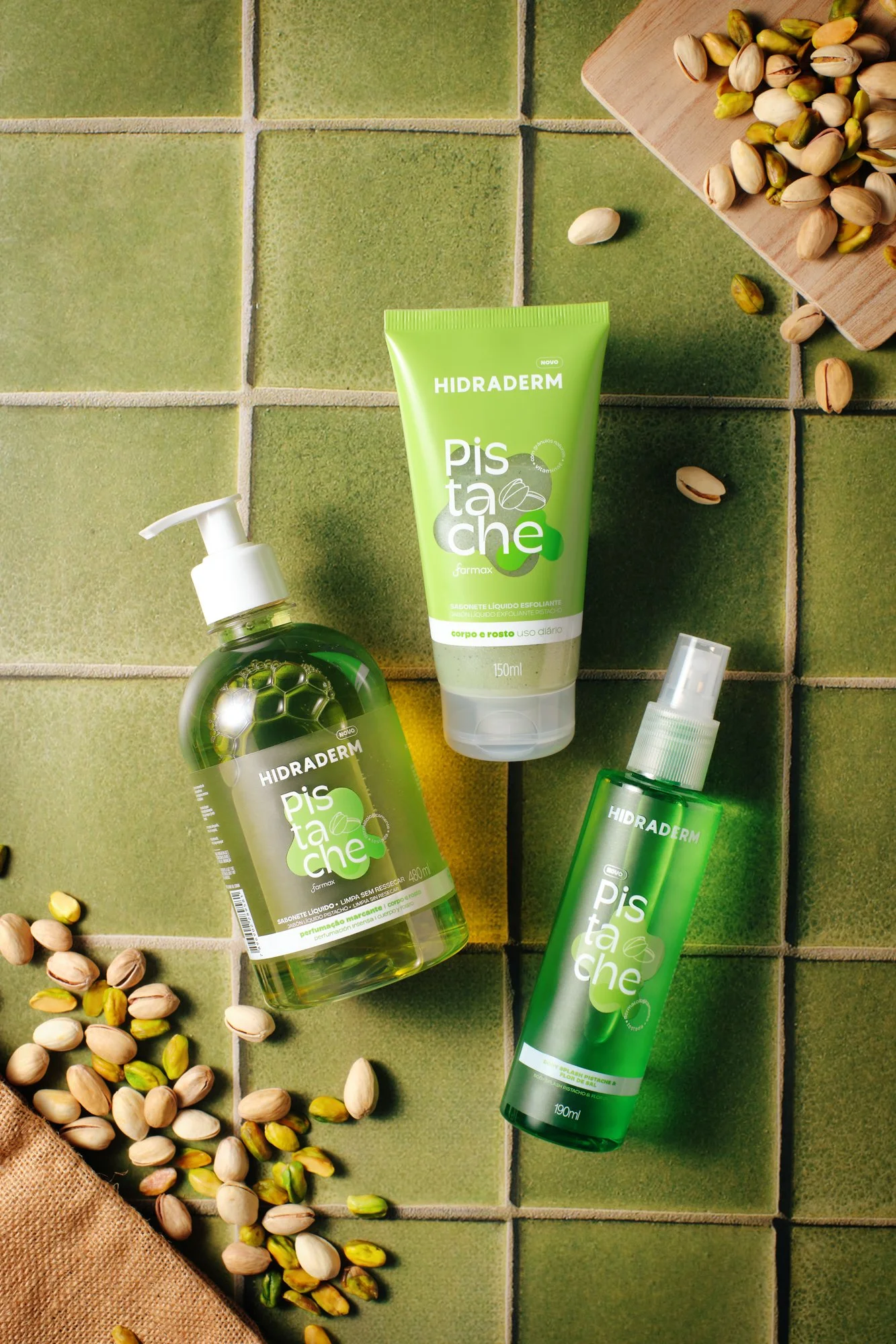 Conjunto de produtos Hidraderm Pista Che de cor verde, incluindo sabonete líquido, creme esfoliante e spray, dispostos sobre azulejos verdes, com pistaches ao redor.