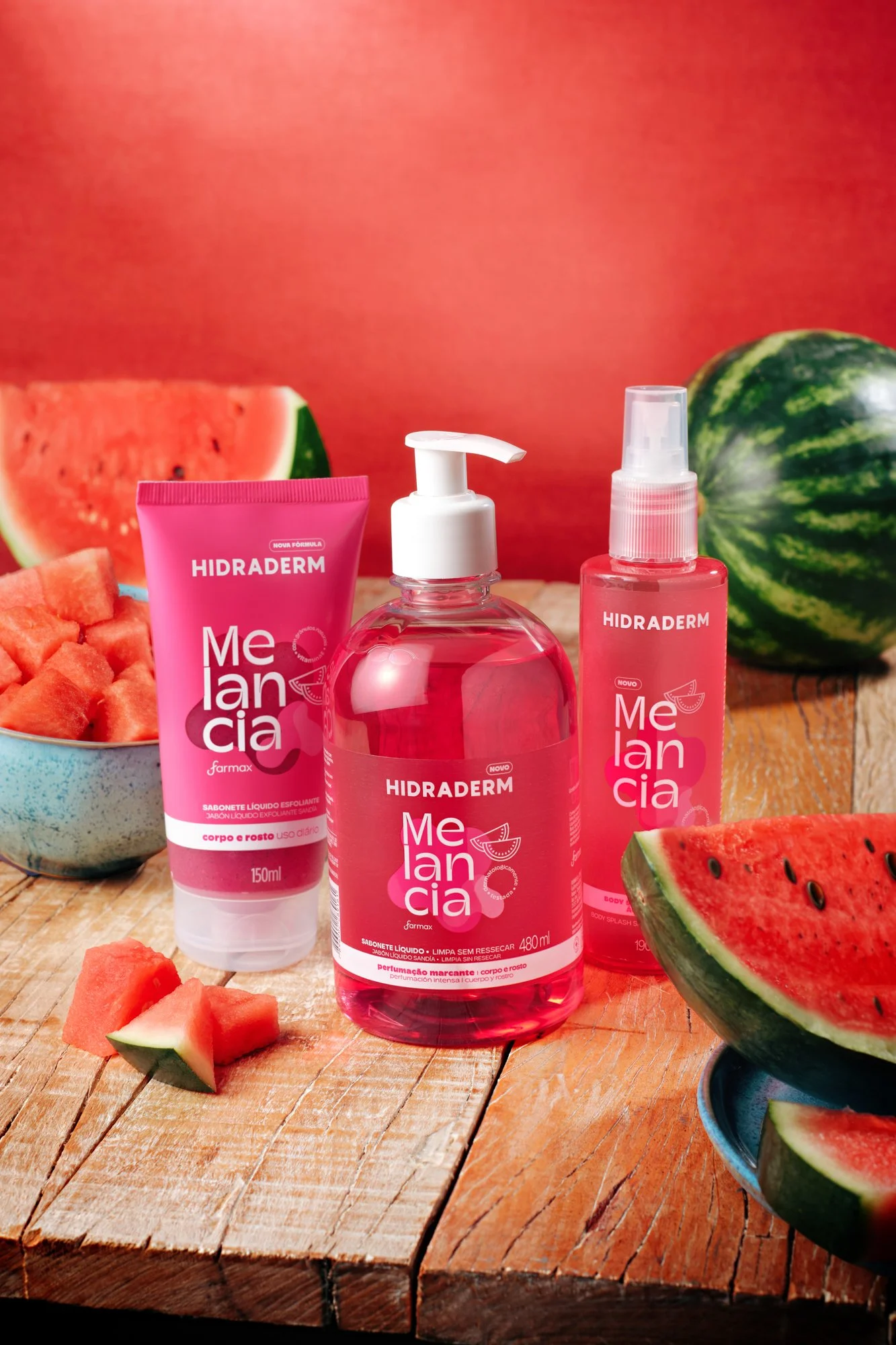 Produtos de cuidados com a pele Melancia Hidradem, incluindo sabonete líquido e spray, dispostos ao lado de fatias e uma melancia inteira, sobre uma mesa de madeira com fundo vermelho.