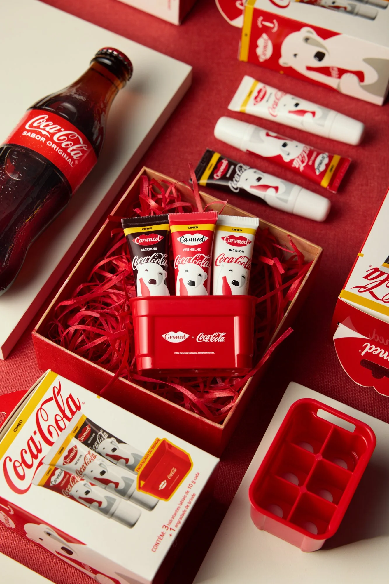 Conjunto de produtos promocionais da Coca-Cola, incluindo tubos de creme com bichinho pinguim e caixinha com compartimentos, além de uma garrafa de Coca-Cola de vidro. Os produtos têm cores vermelha, branca, preta e amarela, com desenhos de um pingui