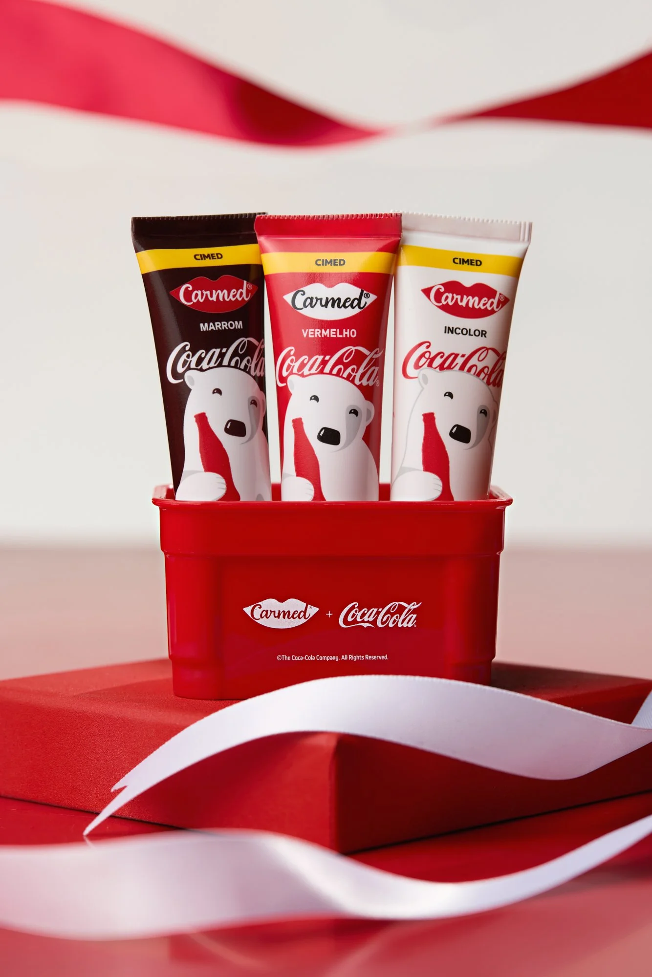 Três tubos de creme intercalando cores marrom, vermelho e branco, com personagem de urso segurando uma garrafa de Coca-Cola, em uma caixa vermelha com logotipos da Carmed e Coca-Cola.