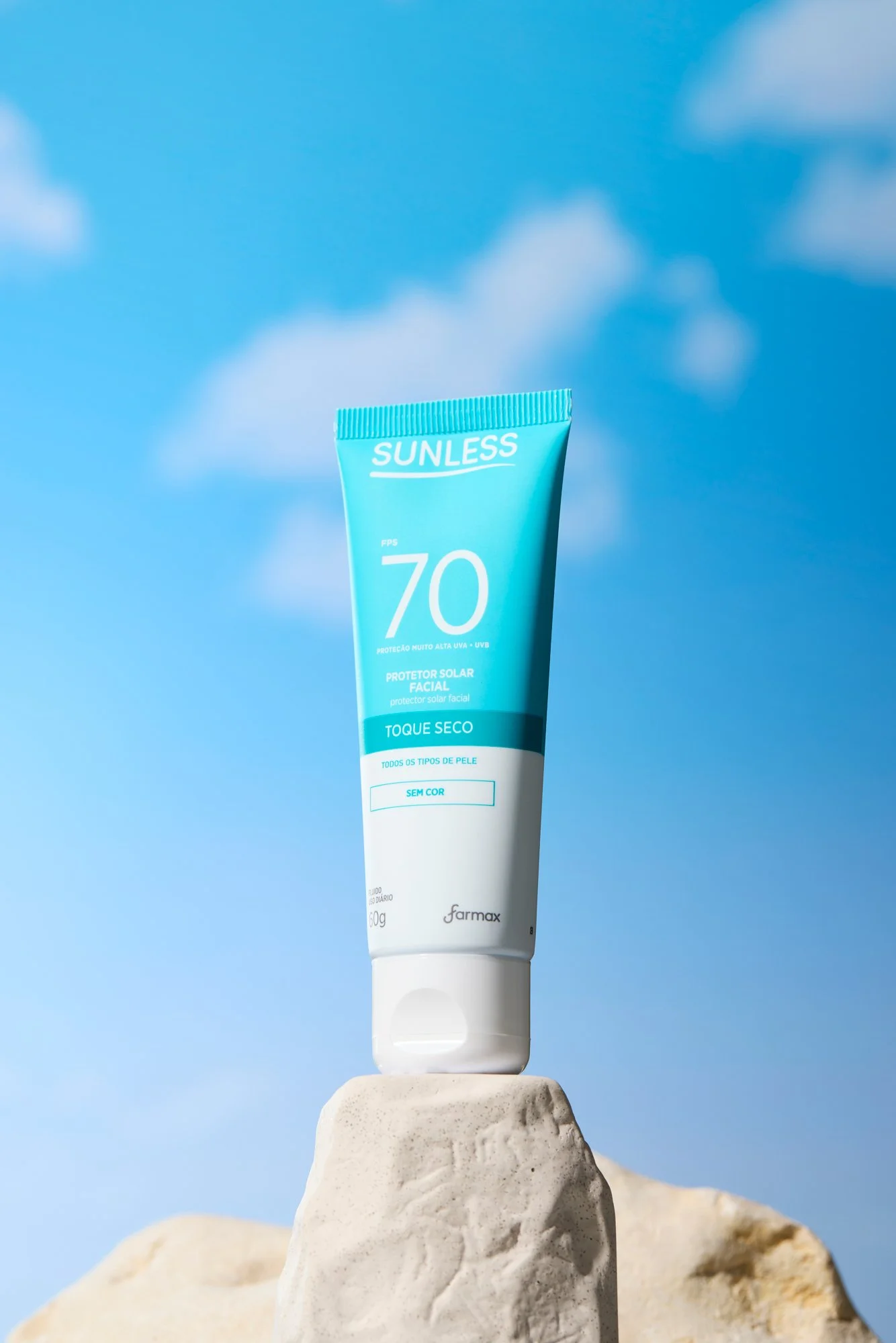 Protetor solar facial Sunless FPS 70 em embalagem azul e branco, apoiado sobre uma pedra, com céu azul ao fundo