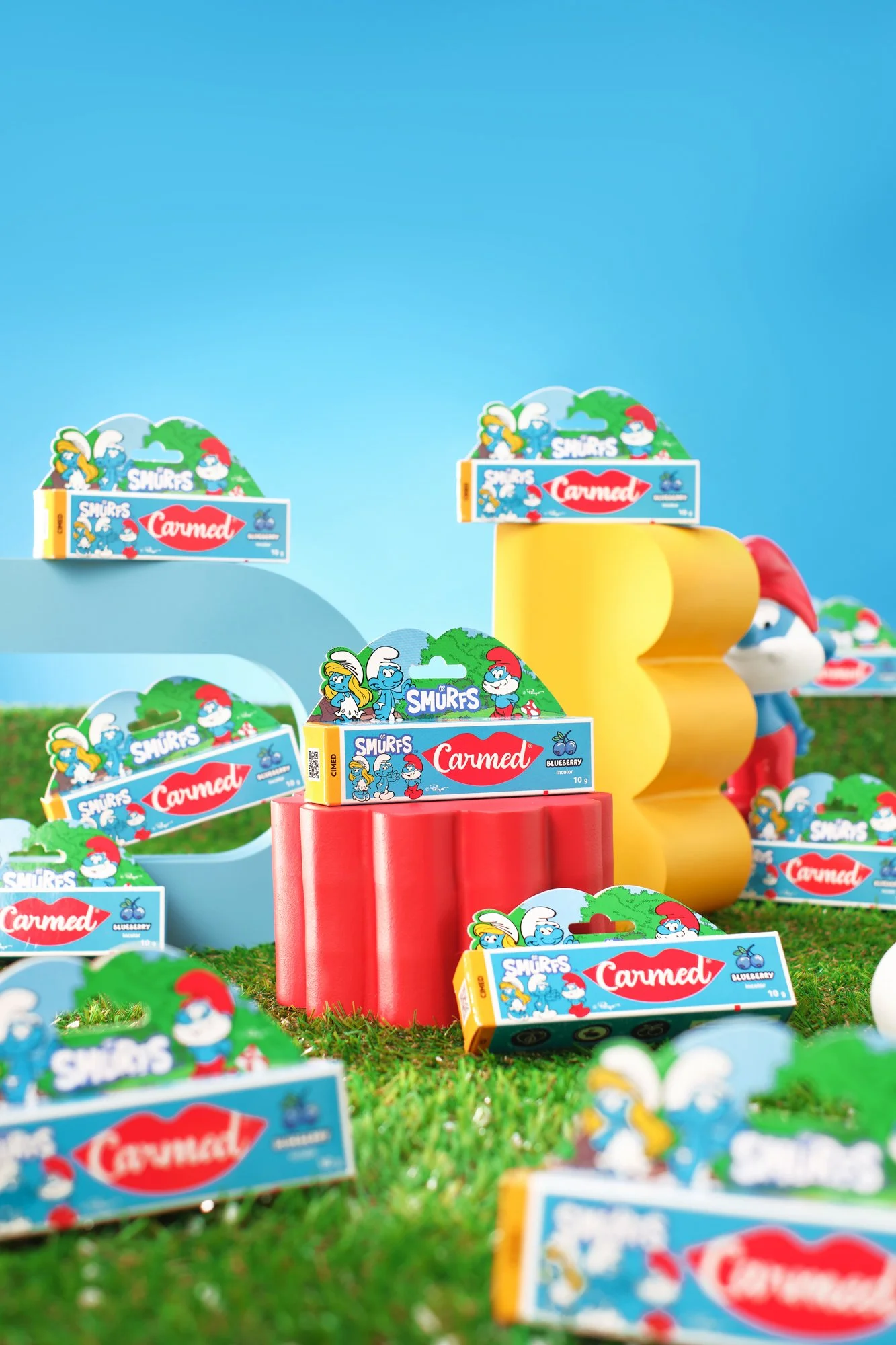 Vários pacotes de bala de goma de personagem dos desenhos animados Smurfs e C summed, com cores verde, azul, vermelho e amarelo, dispostos sobre um tapete de grama artificial, com uma figura de Smurf ao fundo.