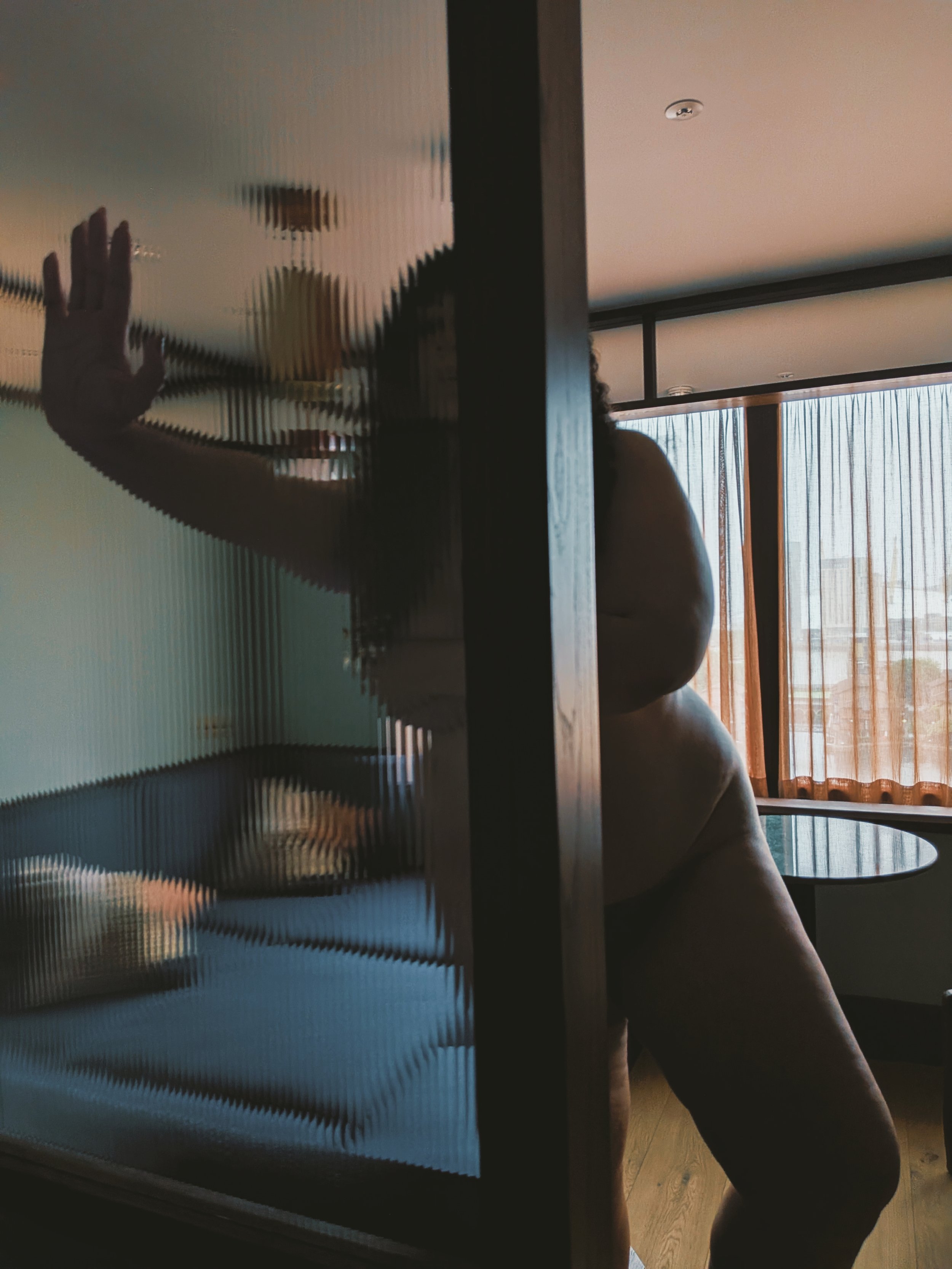 Miss Annie Hope who is a BBW escort from London stands with a hand pressed against the mirror's surface, and a background of large windows with curtains letting in natural light.
