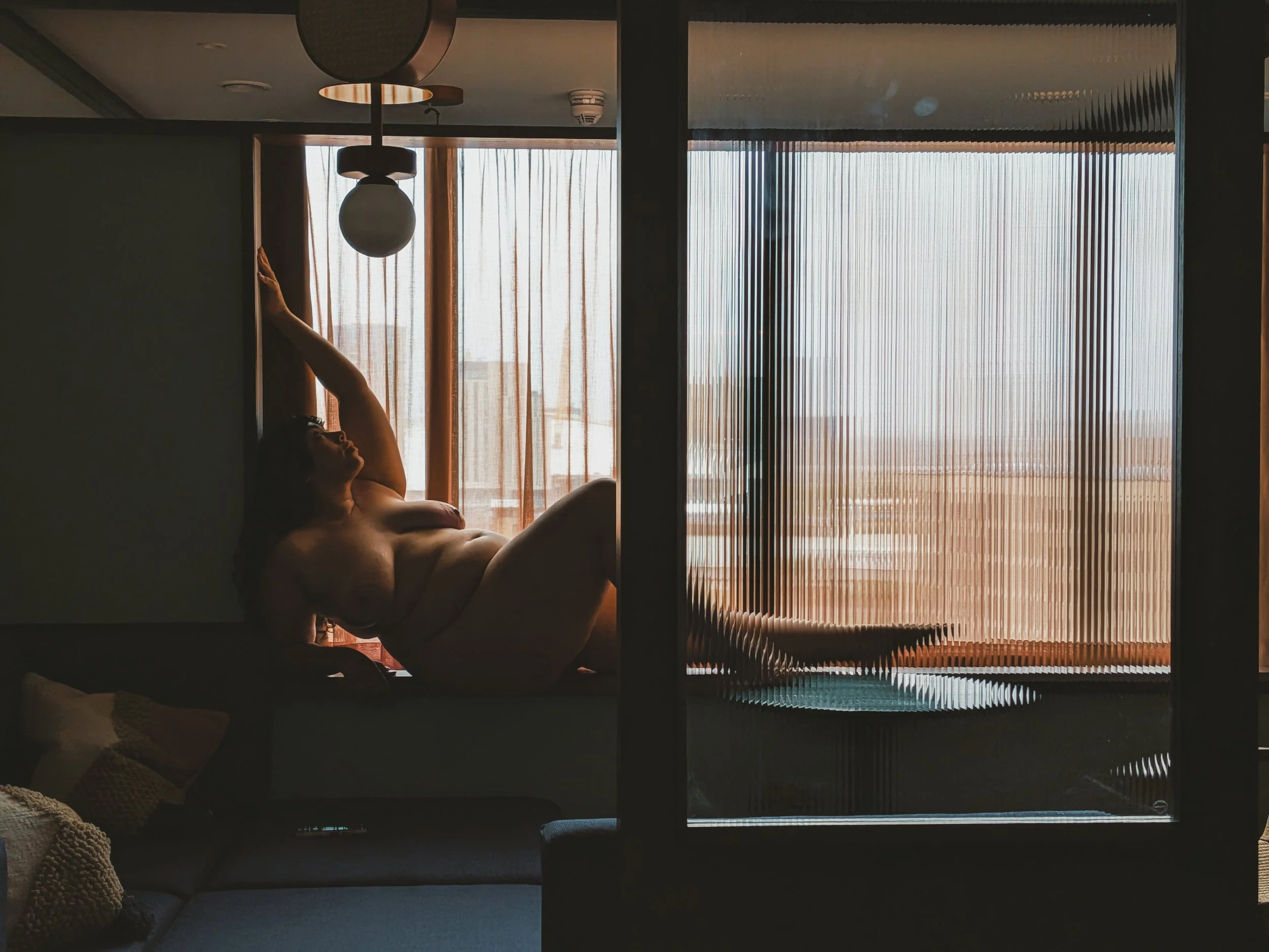 High-class independent BBW escort Miss Annie Hope lies next to a window with sunlight coming through sheer curtains, partially covered by a blanket, in a room seen through a glass door with vertical blinds.
