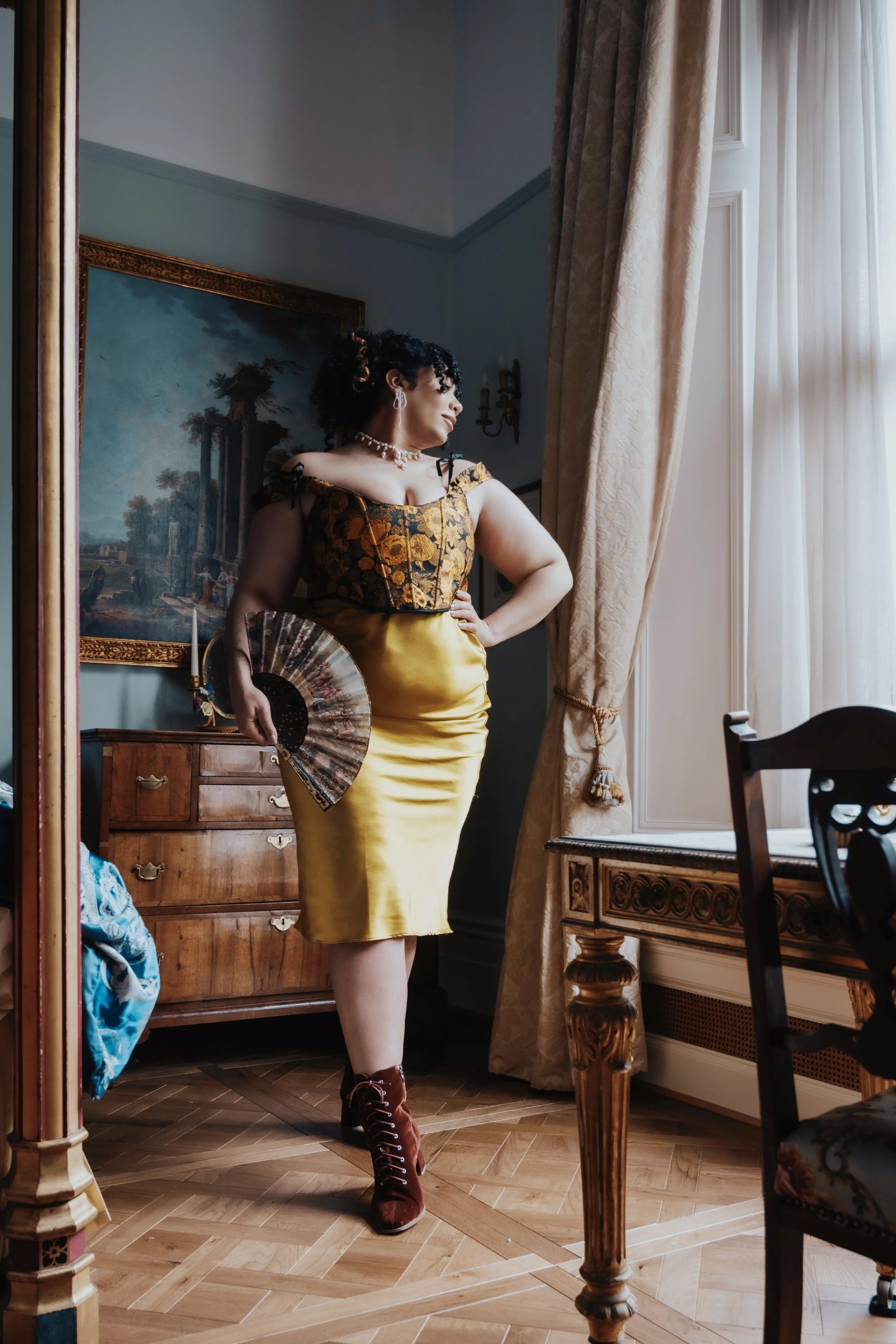 A woman in an elegant yellow dress with floral embroidery on top, standing in a decorated room by a window with heavy curtains, holding a fan, wearing jewelry and lace-up boots, looking out the window.