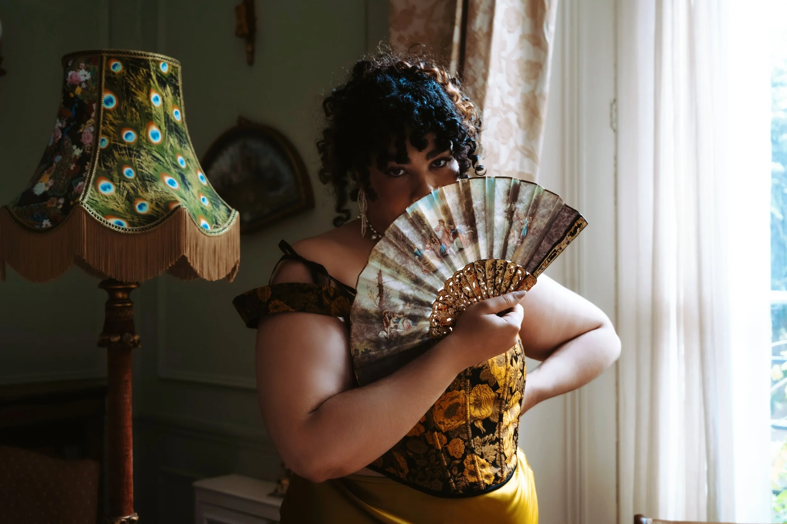 Miss Annie hope BBW escort from London has curly dark hair and dark eye makeup holding a decorative fan in front of her face. There is an ornate, peacock-patterned lampshade on a lamp beside her.