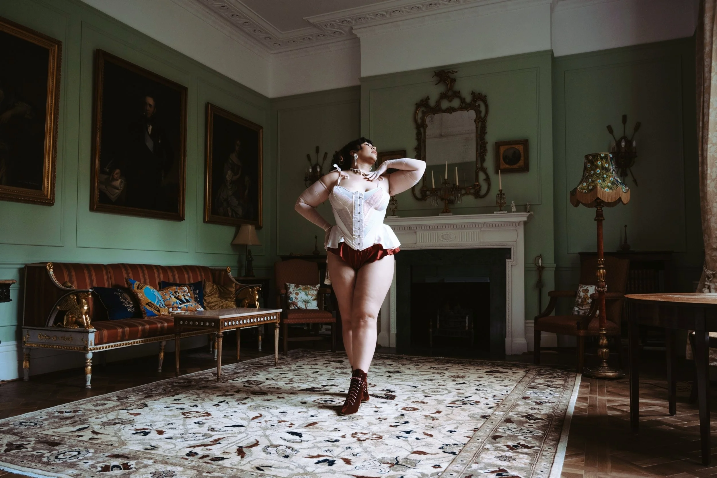 Miss Annie Hope London curvy escort is in vintage lingerie and high-heeled boots standing in an elegant, old-fashioned living room with green walls, ornate decorations, and classic furniture.