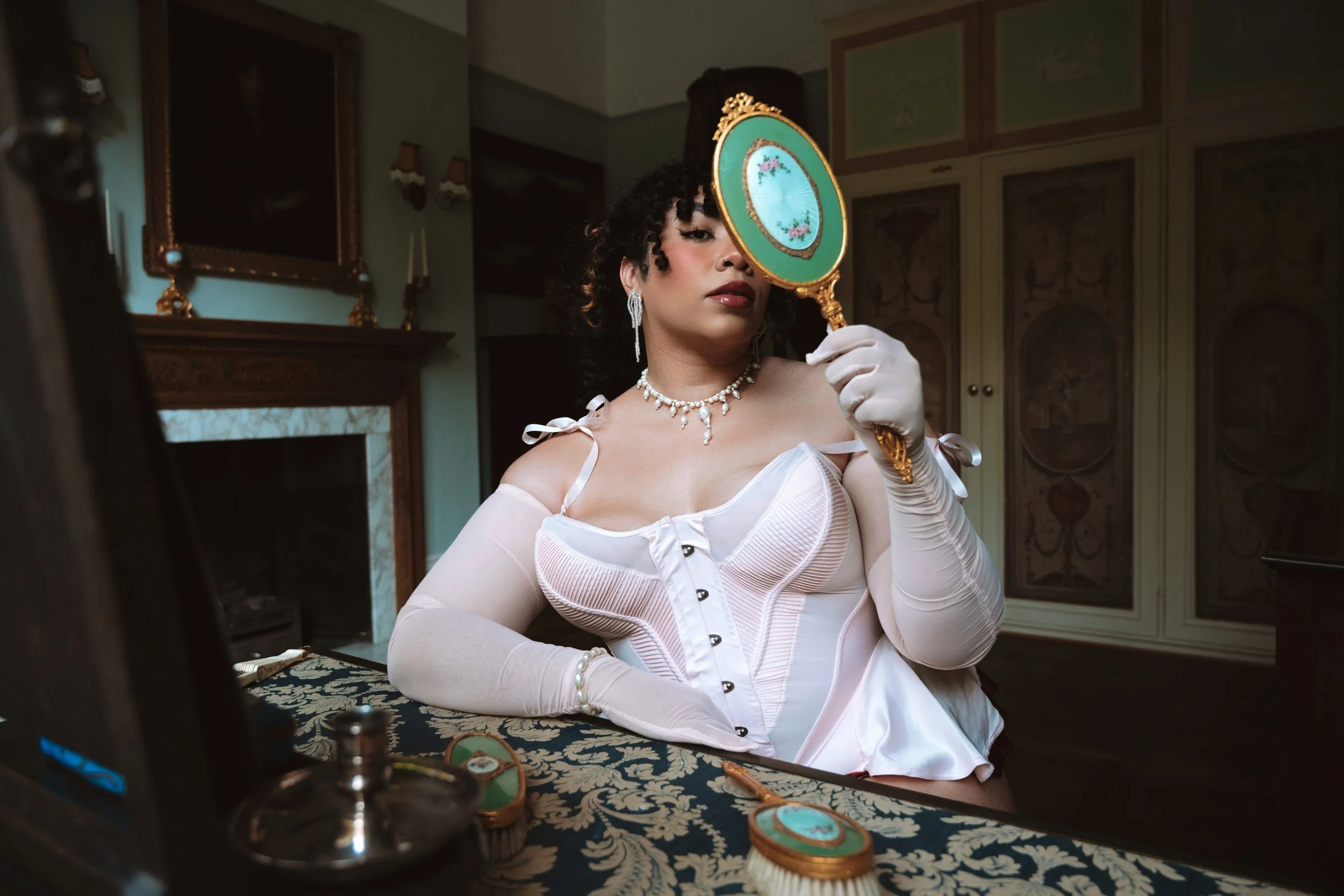 Miss Annie Hope BBW Mistress is dressed in vintage attire wearing jewellery, holding a decorative hand mirror with floral design, sitting at a dressing table with makeup items in a luxurious room.