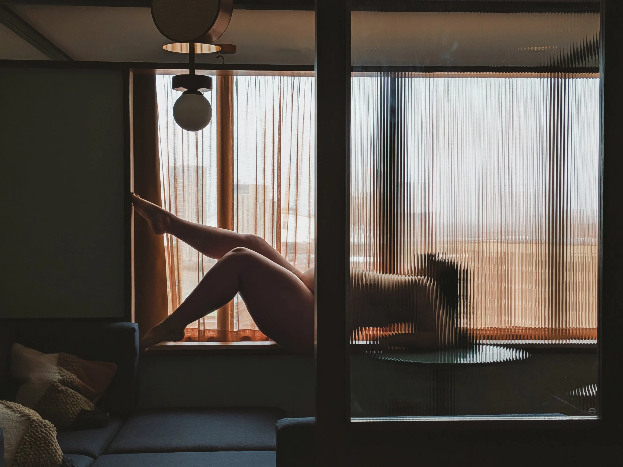 Miss Annie Hope, London BBW escort lays by the window, legs stretched out, with sunlight streaming through sheer curtains, seen through a glass partition in a dimly lit room.