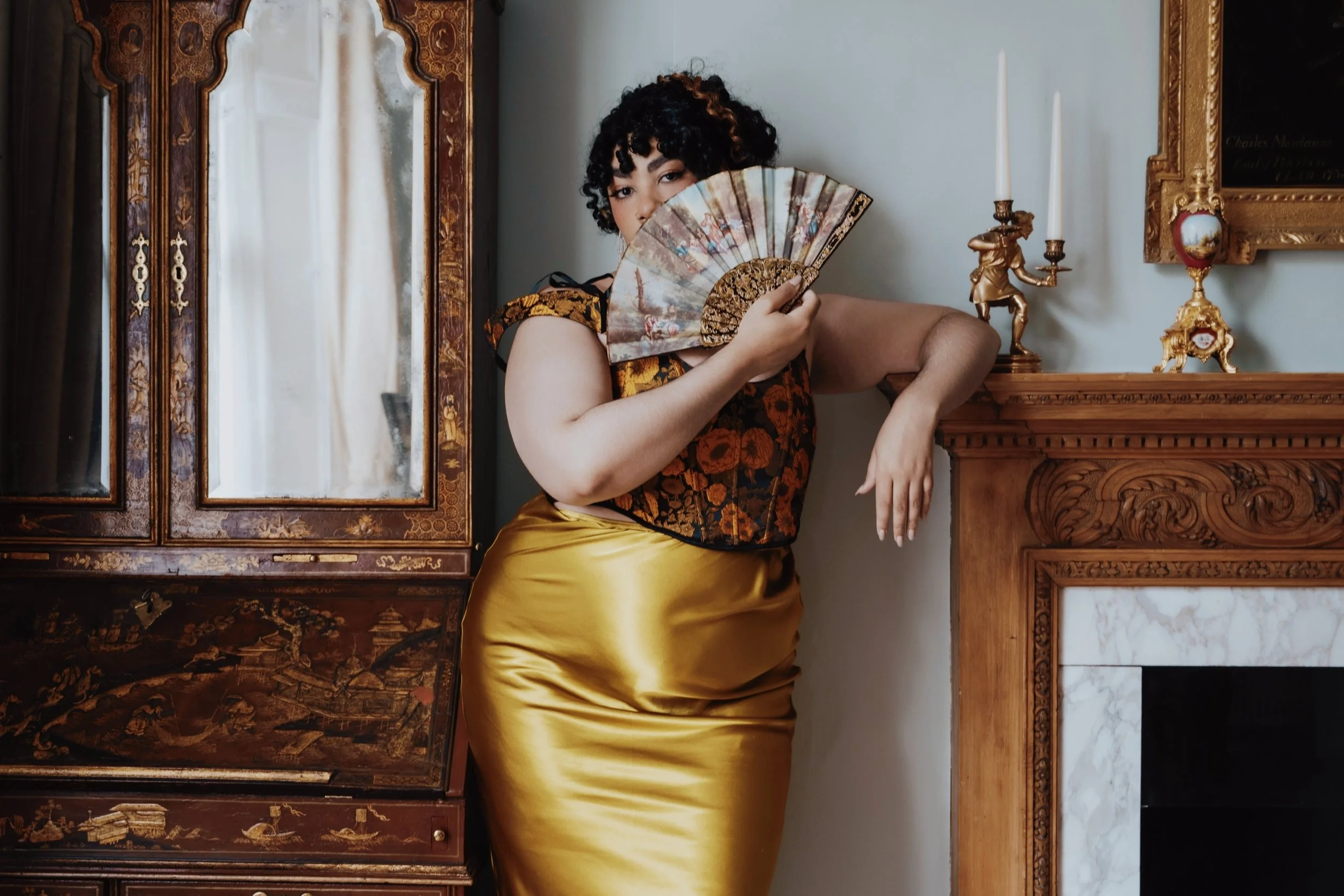 Miss Annie Hope BBW escort and Mommy Domme from London has curly hair and holds a decorative hand fan partially covering her face. She is dressed in a gold corset and a shiny gold skirt, standing beside an ornate wooden cabinet and a fireplace.