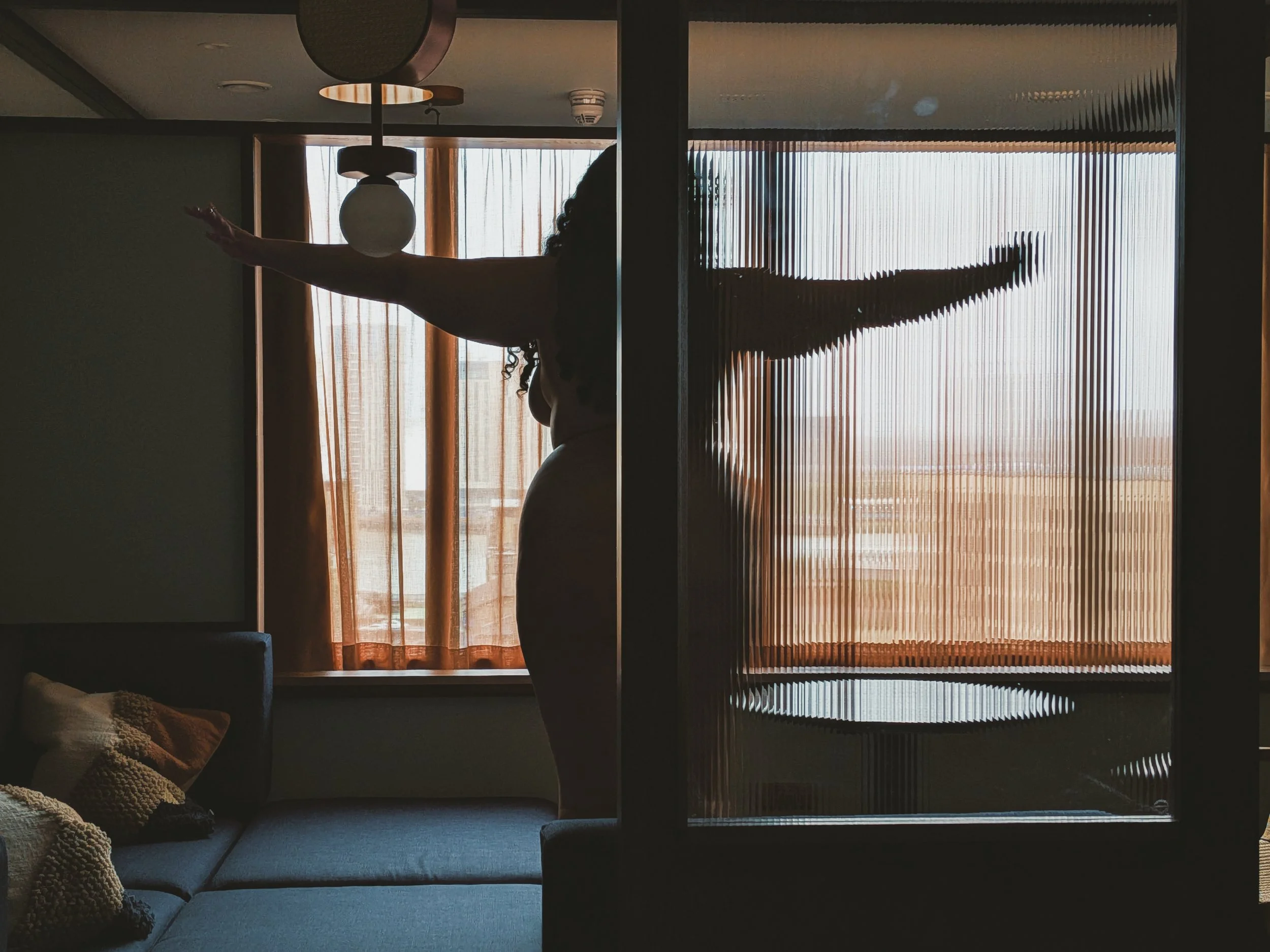 A silhouette of Independent escort Miss Annie Hope with curly hair standing in front of a large window with vertical blinds, arms extended outward. She is naked.