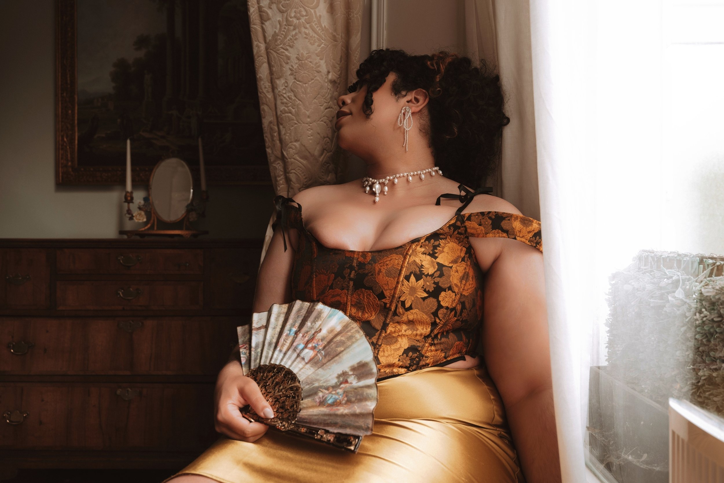 Miss Annie Hope BBW Mommy Domme and ABDL escort has curly hair and wears elegant jewellery, including earrings and a pearl necklace, is sitting near a window with sheer curtains, holding a decorative fan