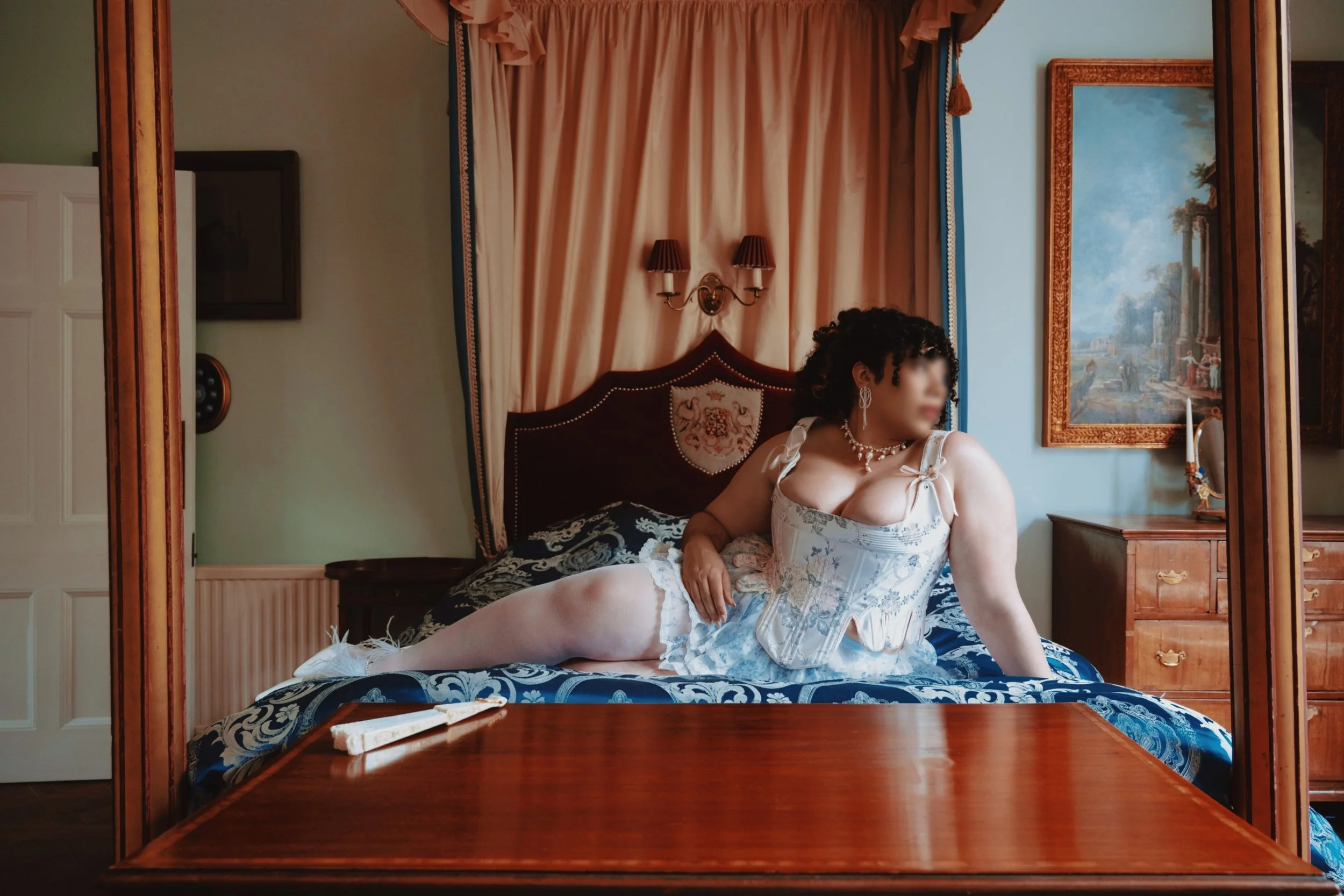 Miss Annie Hope, Luxury independent escort and curvy mommy domme sits on an ornate bed wearing a white corset