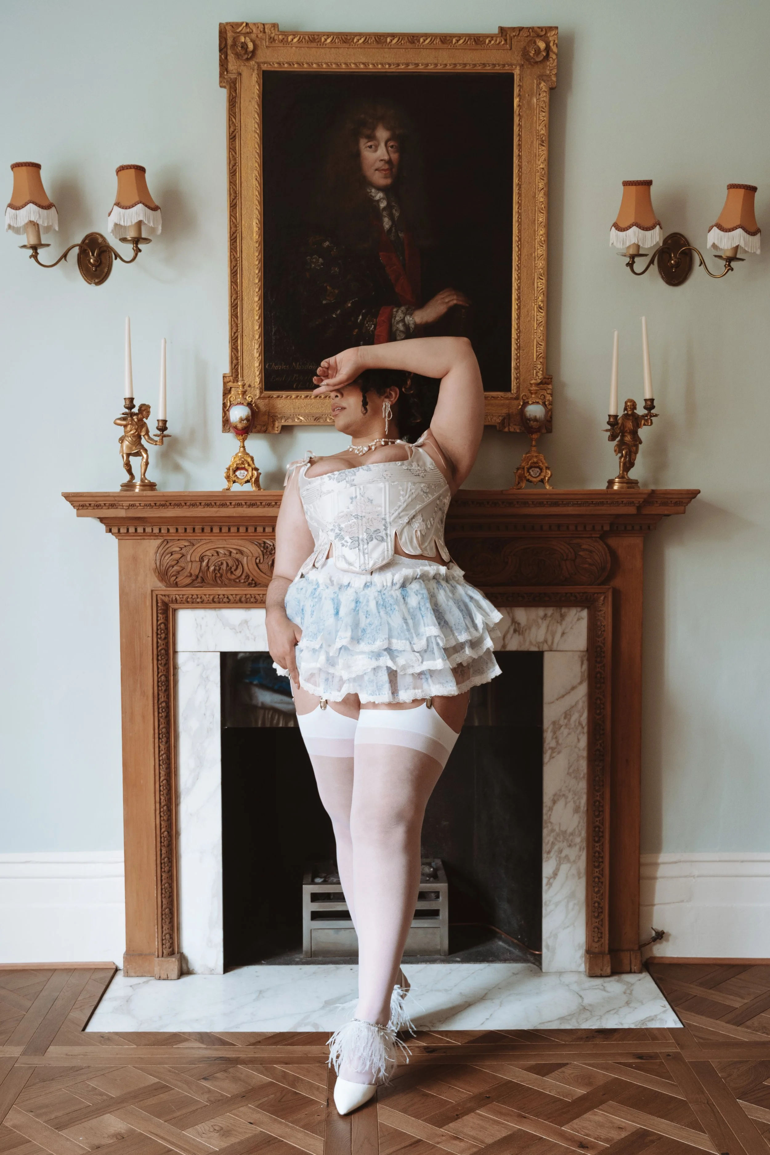 Gentle Mommy Domme and BBW kinky companion escort Miss Annie Hope leans against a fireplace wearing a white corset and baby blue frilly bloomers