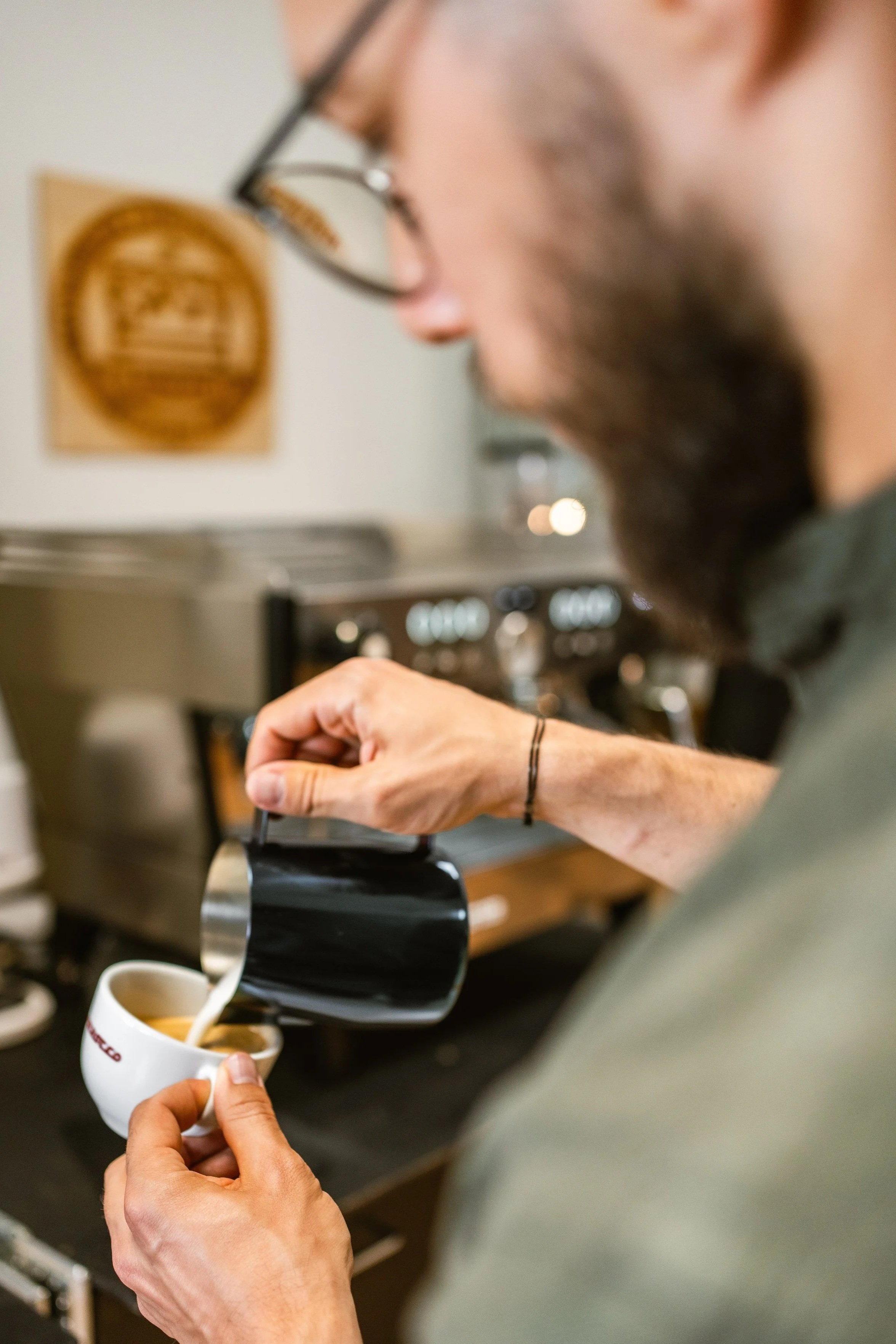 Ein Barista gießt Latte Art in eine Tasse und zeichnet eine Tulpe auf einen Cappuccino. Im Hintergrund eine Espressomaschine.