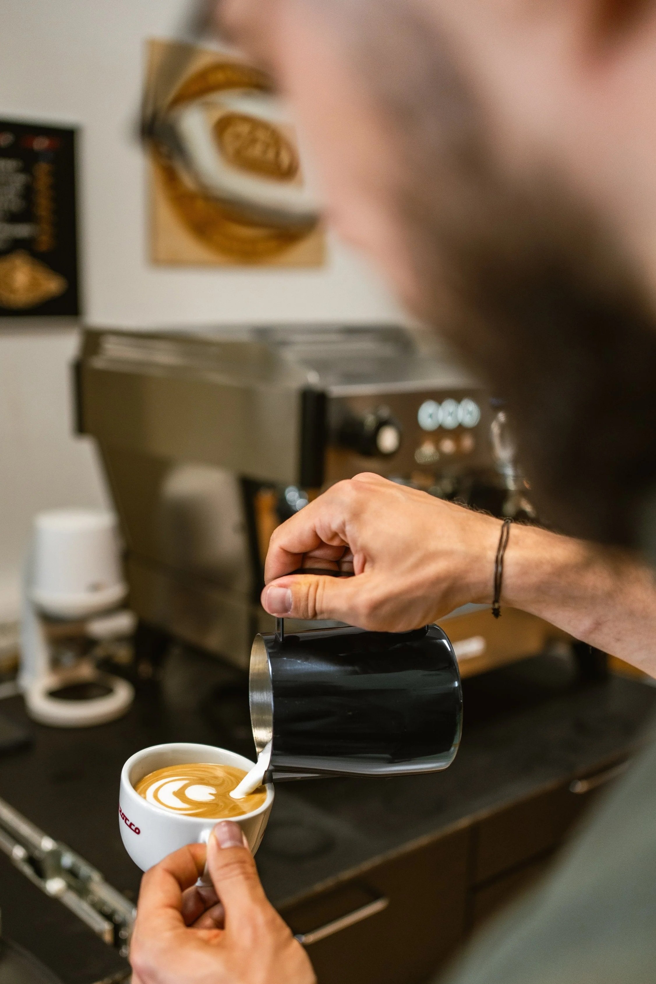 Ein Barista gießt Latte Art in eine Tasse und zeichnet eine Tulpe auf einen Cappuccino. Im Hintergrund eine Espressomaschine.