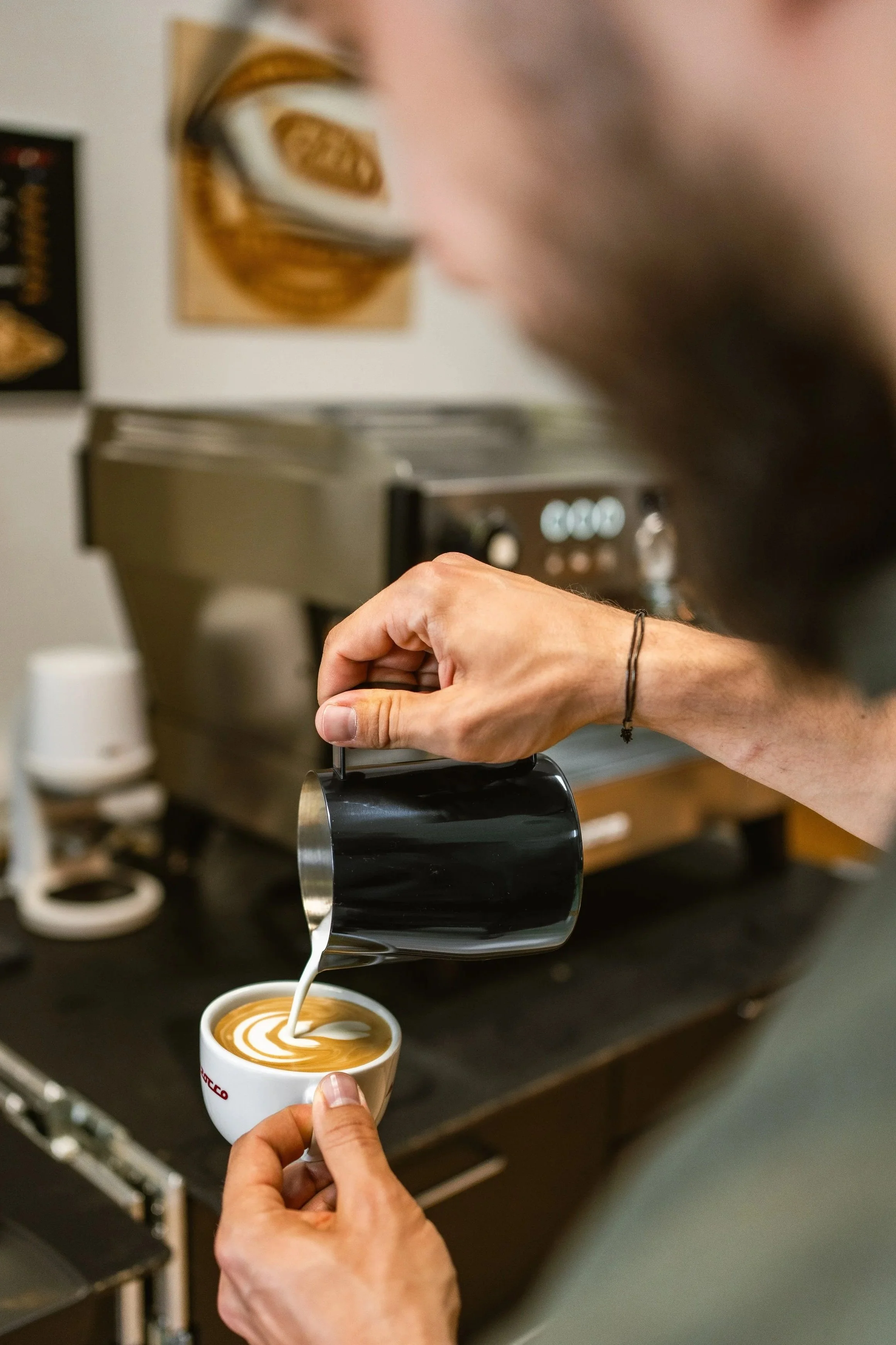 Ein Barista gießt Latte Art in eine Tasse und zeichnet eine Tulpe auf einen Cappuccino. Im Hintergrund eine Espressomaschine.