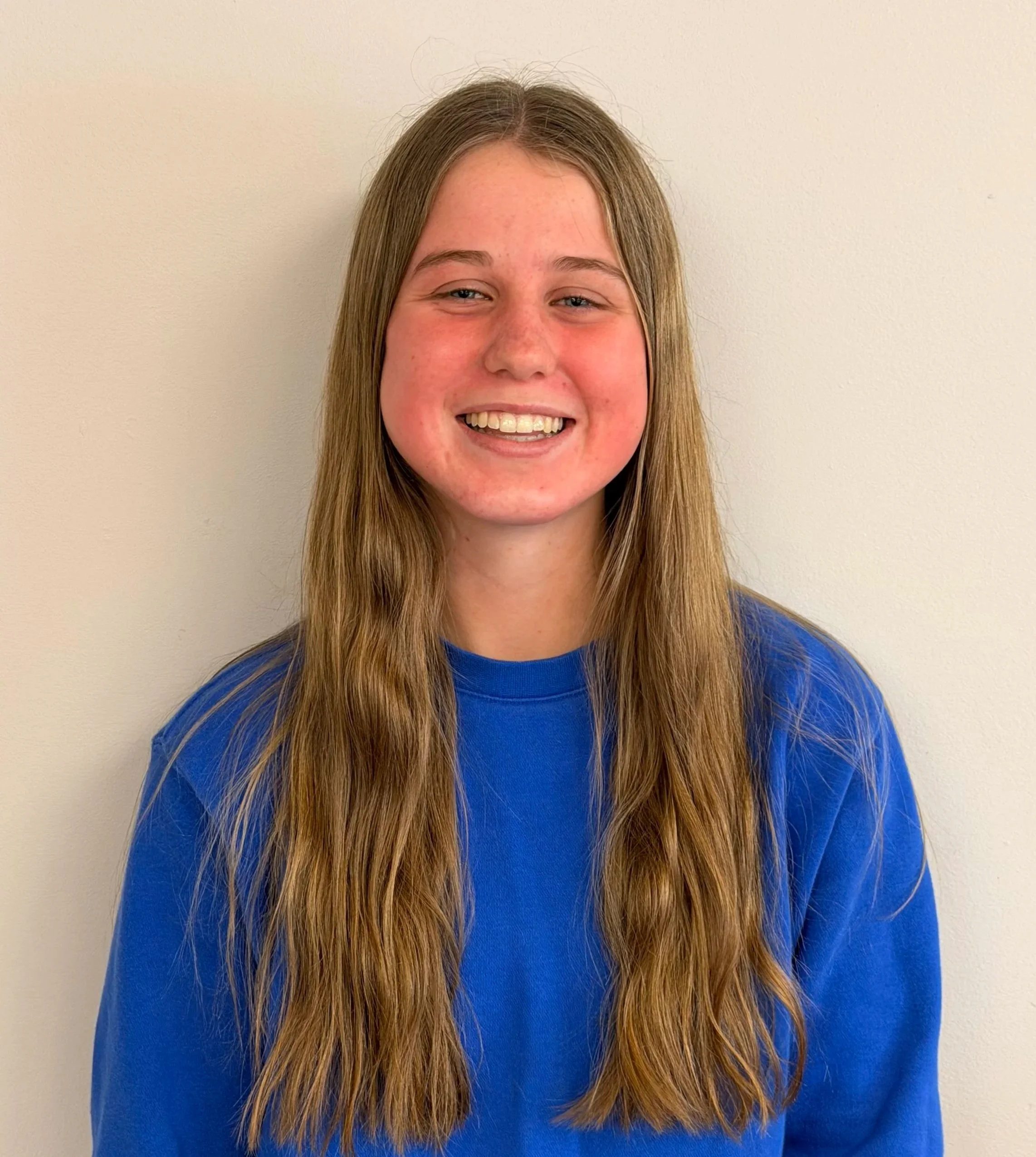 Portrait of a young woman with long, straight, light brown hair, smiling and wearing a blue shirt, standing against a plain, light-colored background.