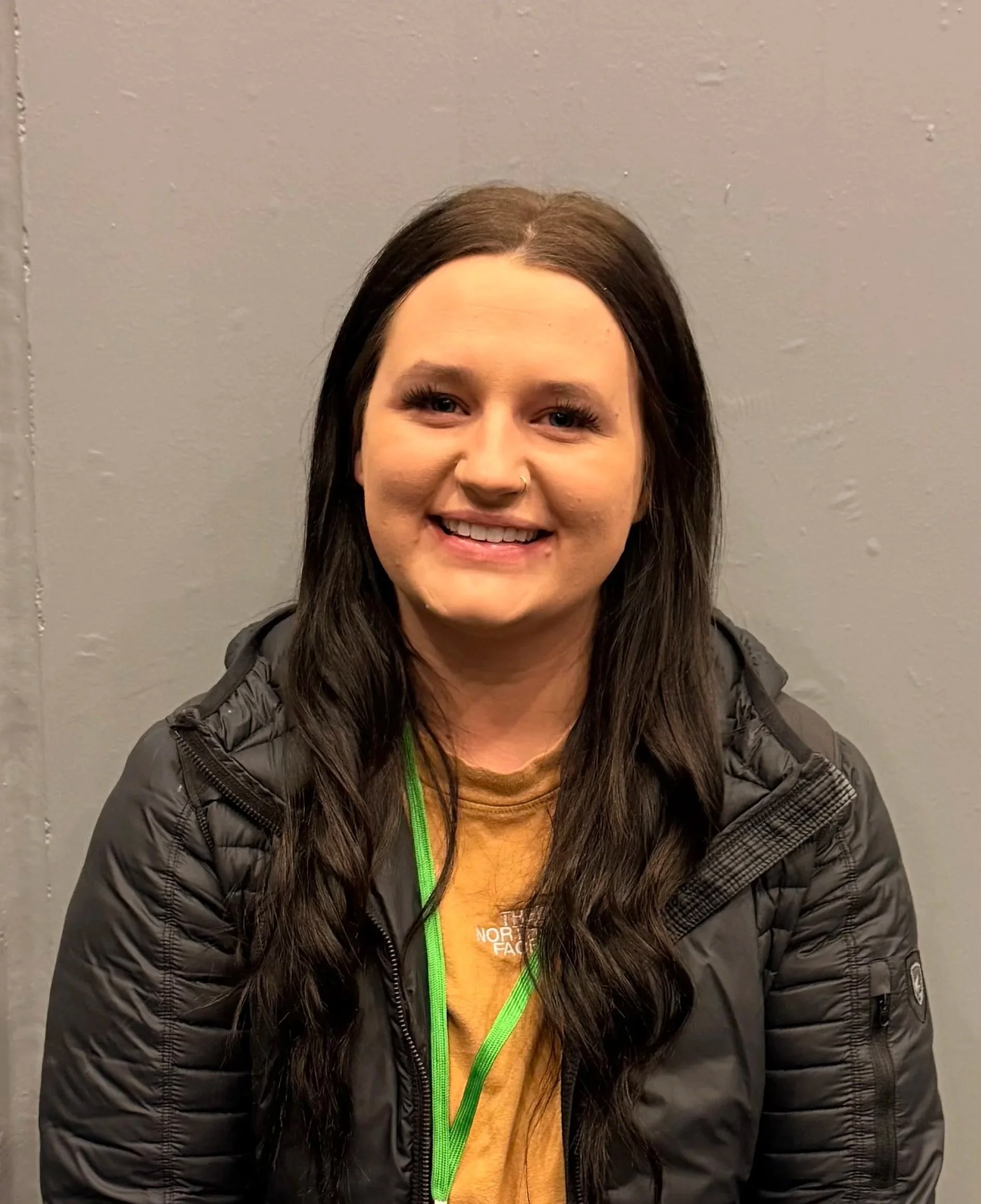 A woman with long dark hair, fair skin, and a nose piercing, smiling, wearing a yellow shirt, a black jacket, and a green lanyard, standing in front of a plain gray wall.