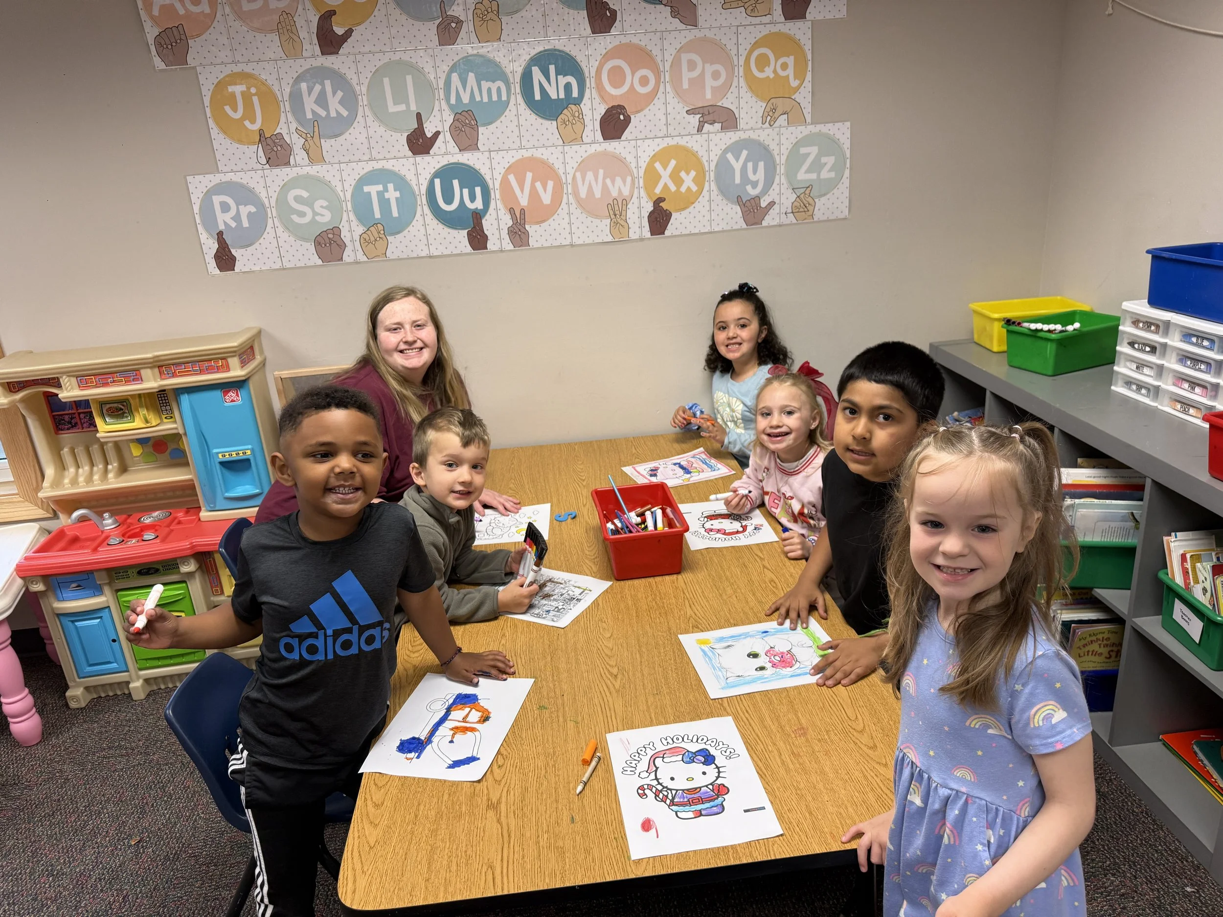 A group of children and a woman smiling at a table in a classroom, engaging in coloring activities with drawings of Hello Kitty and other characters. The classroom has educational posters of the alphabet and various toys and art supplies.