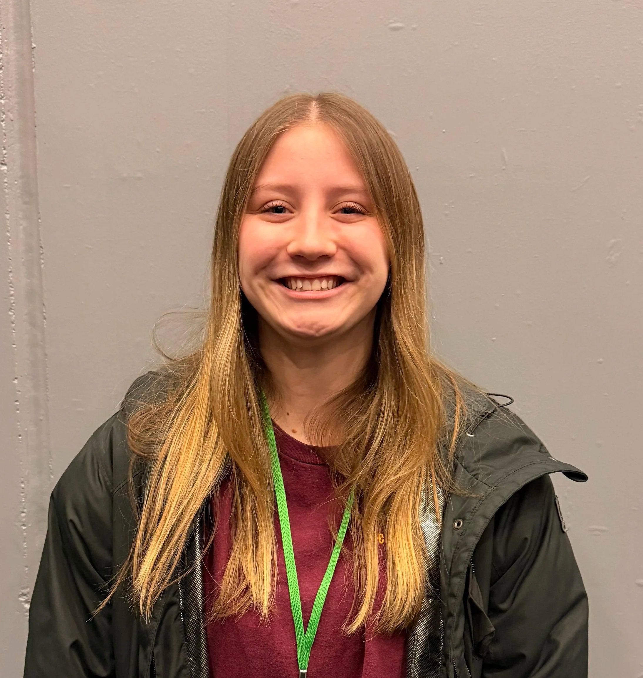 Close-up of a smiling young girl with long blonde hair, wearing a dark jacket over a maroon shirt, standing against a plain gray wall.