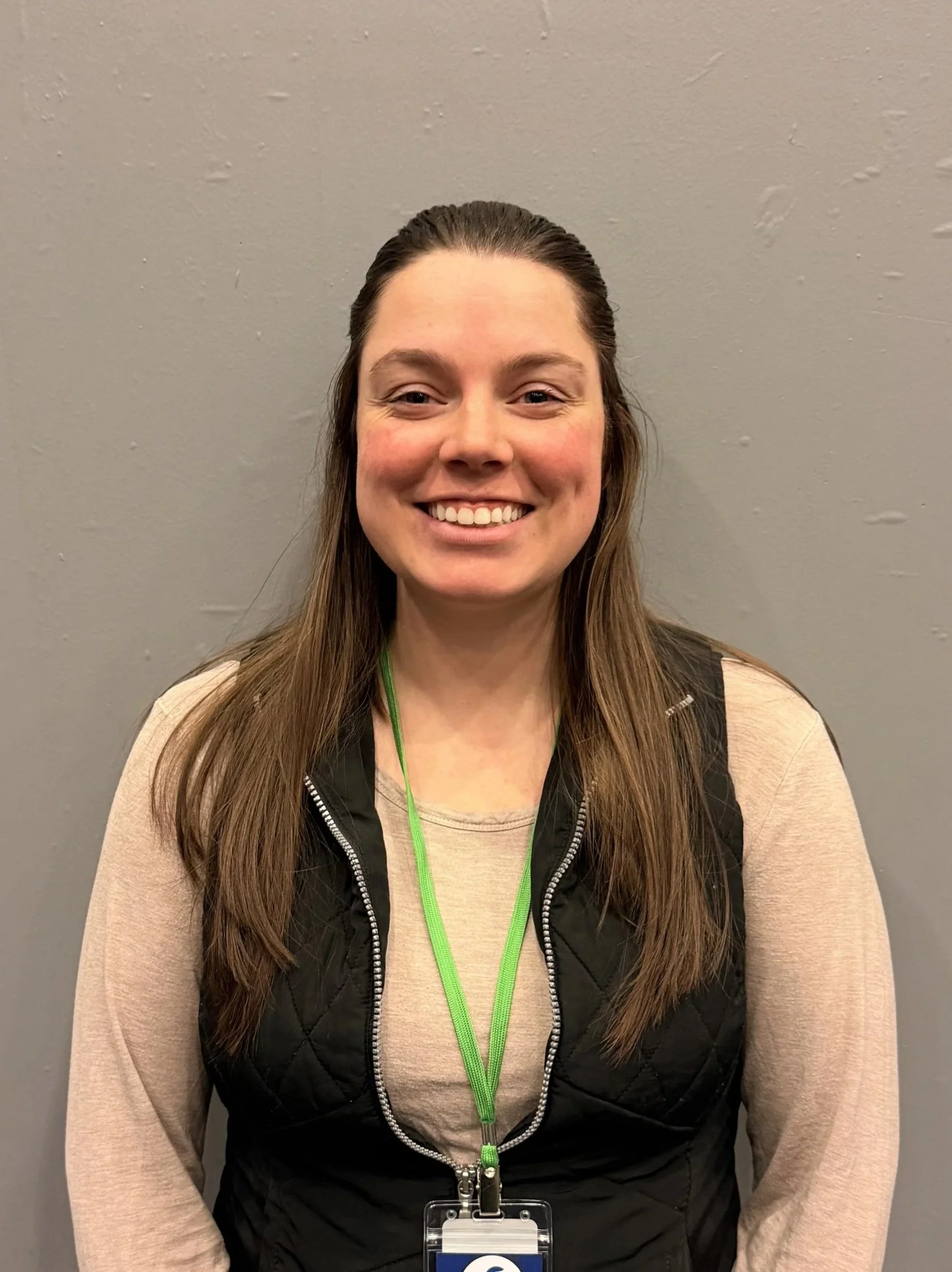 Smiling woman with long brown hair, wearing a beige long-sleeve shirt, black quilted vest, and a green lanyard with a badge, standing against a plain gray wall.