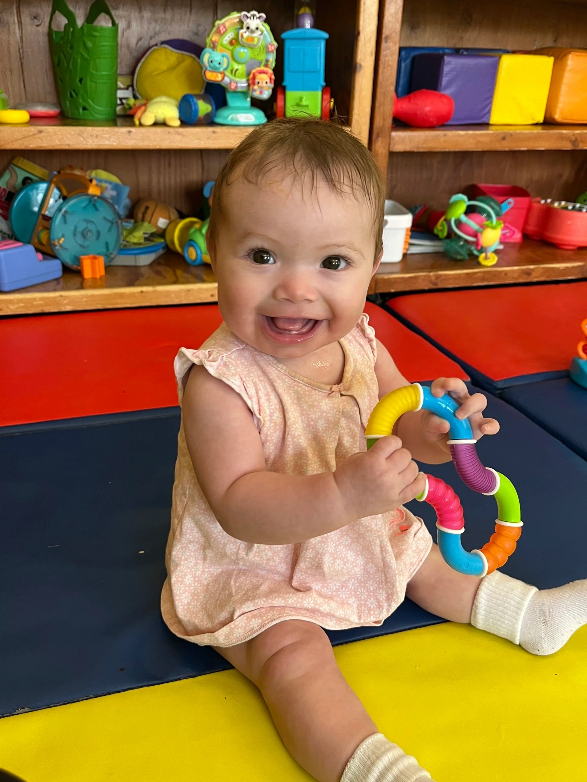 A smiling baby girl sitting on a multicolored play mat, holding a colorful toy ring, with toy shelves filled with various toys in the background.