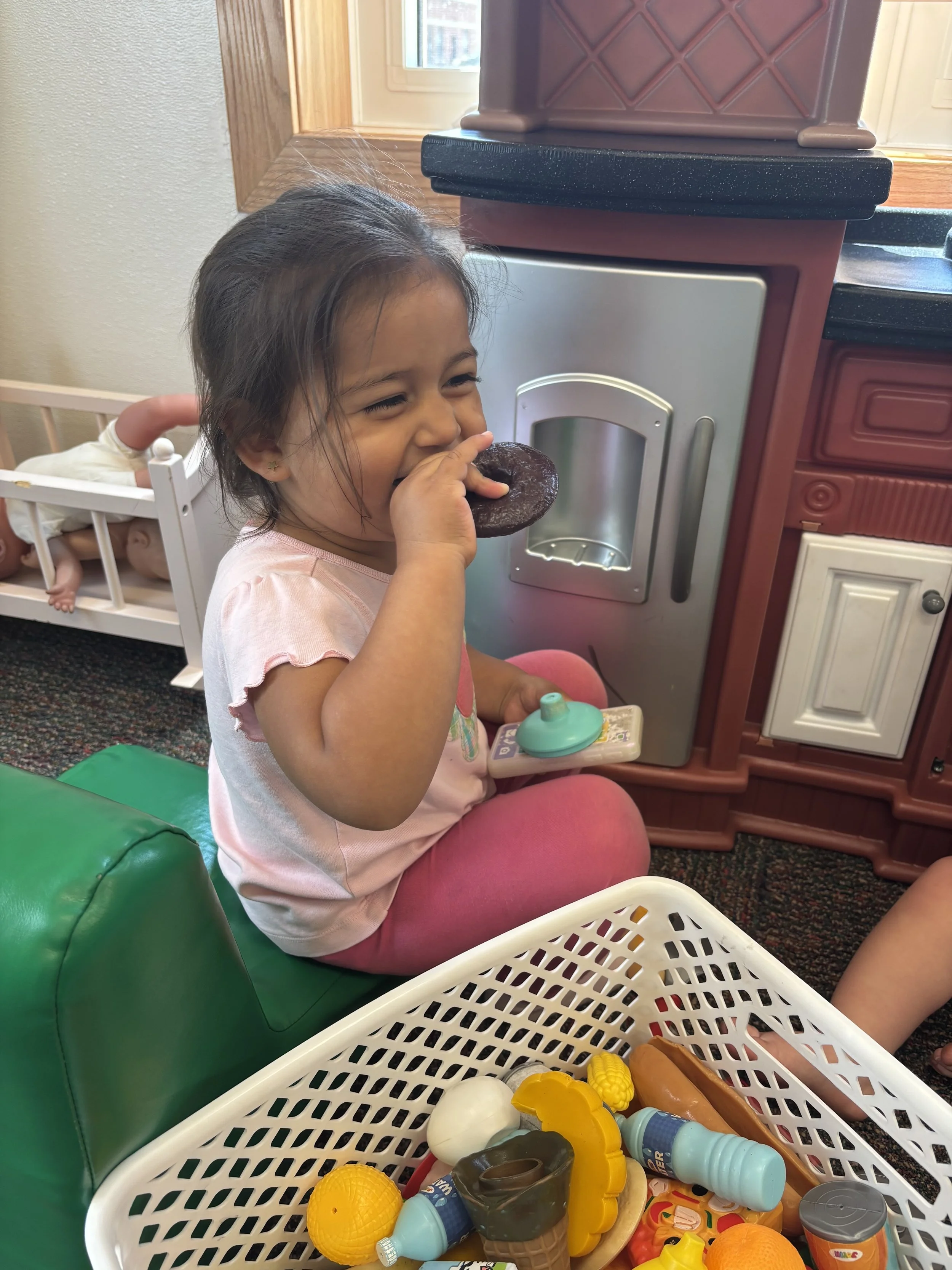 A young girl with dark hair seated on a green chair, holding a toy donut near her mouth and smiling. Behind her, there is a doll in a crib, and in the foreground, a white basket filled with various plastic food toys, including fruits, vegetables, and bottles.