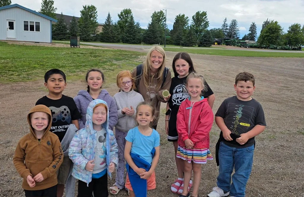 A group of children and an adult woman smiling outdoors on a cloudy day, standing on a gravel area with trees and a building in the background.