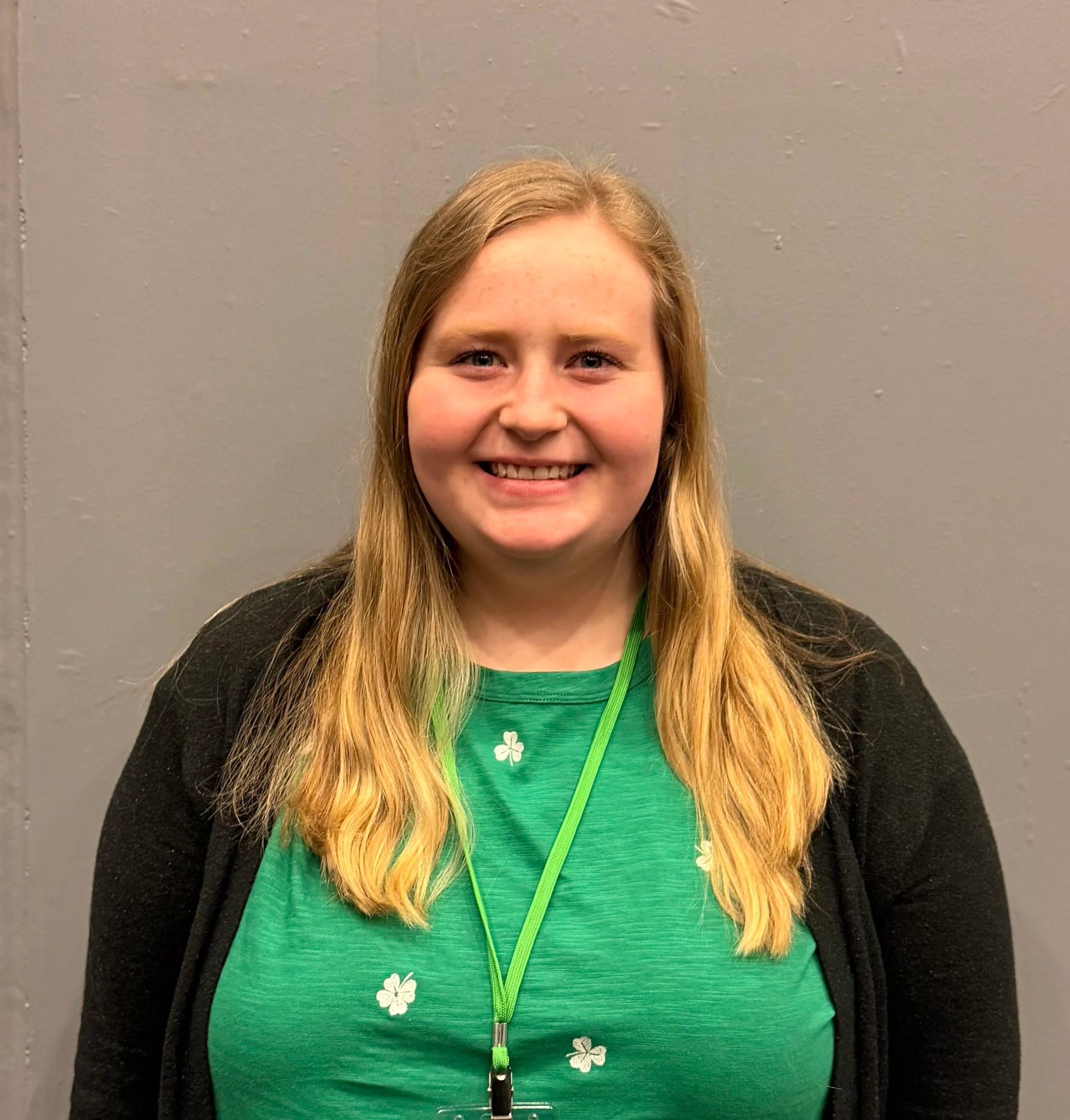 Close-up of a young woman with long blonde hair, smiling, wearing a green shirt with white shamrock patterns and a black jacket, standing in front of a plain gray wall.