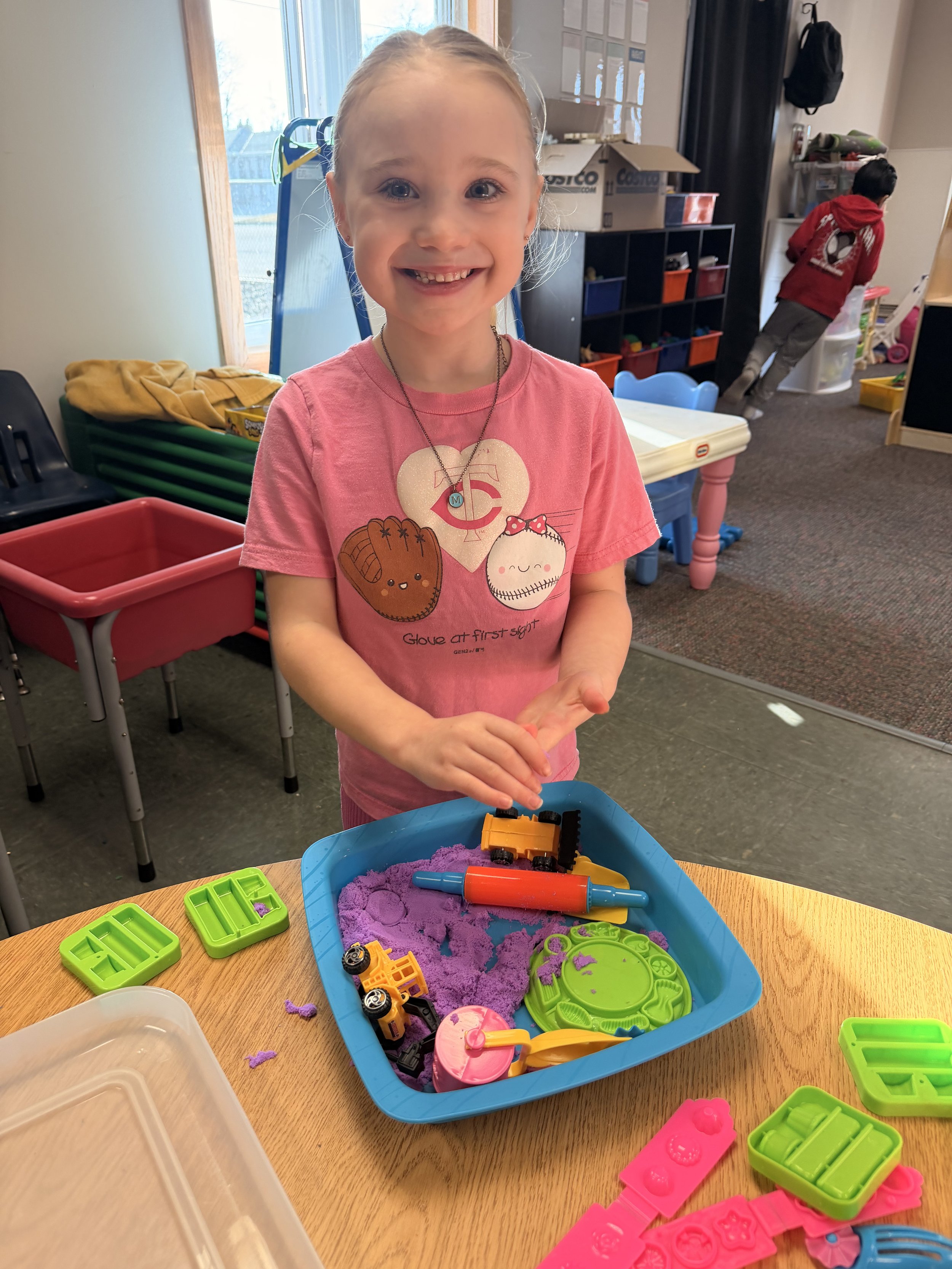 A young girl in a pink shirt smiling at the camera while playing with colored sand and toy molds at a table. In the background, a boy in a red hoodie is leaning over a shelf in a classroom or playroom with toys and storage containers.