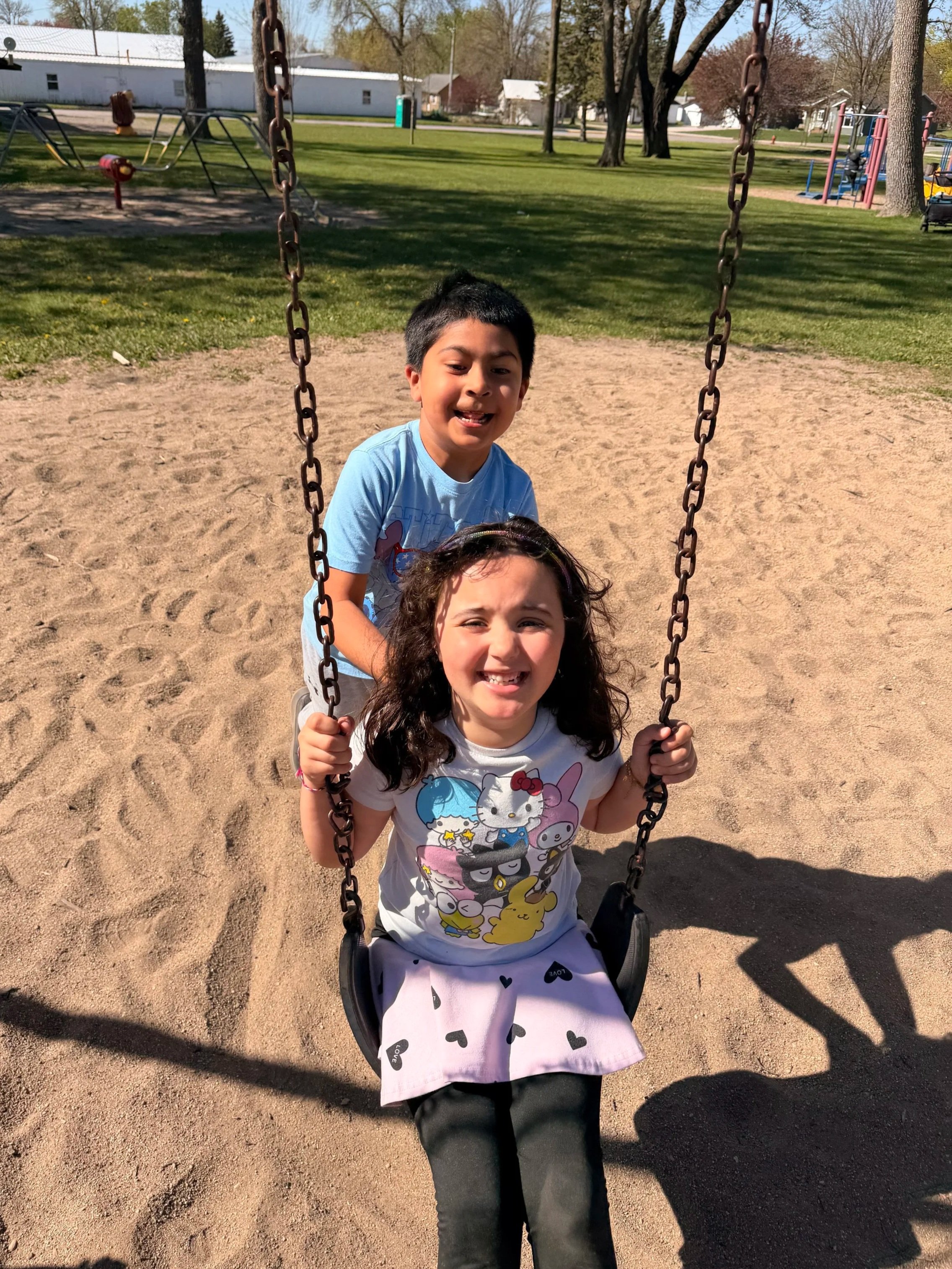 Two children, a girl and a boy, playing on a swing set in a park on a sunny day. The girl is sitting on the swing, smiling at the camera, and the boy is standing behind her, also smiling. In the background, there are trees, houses, and playground equipment.