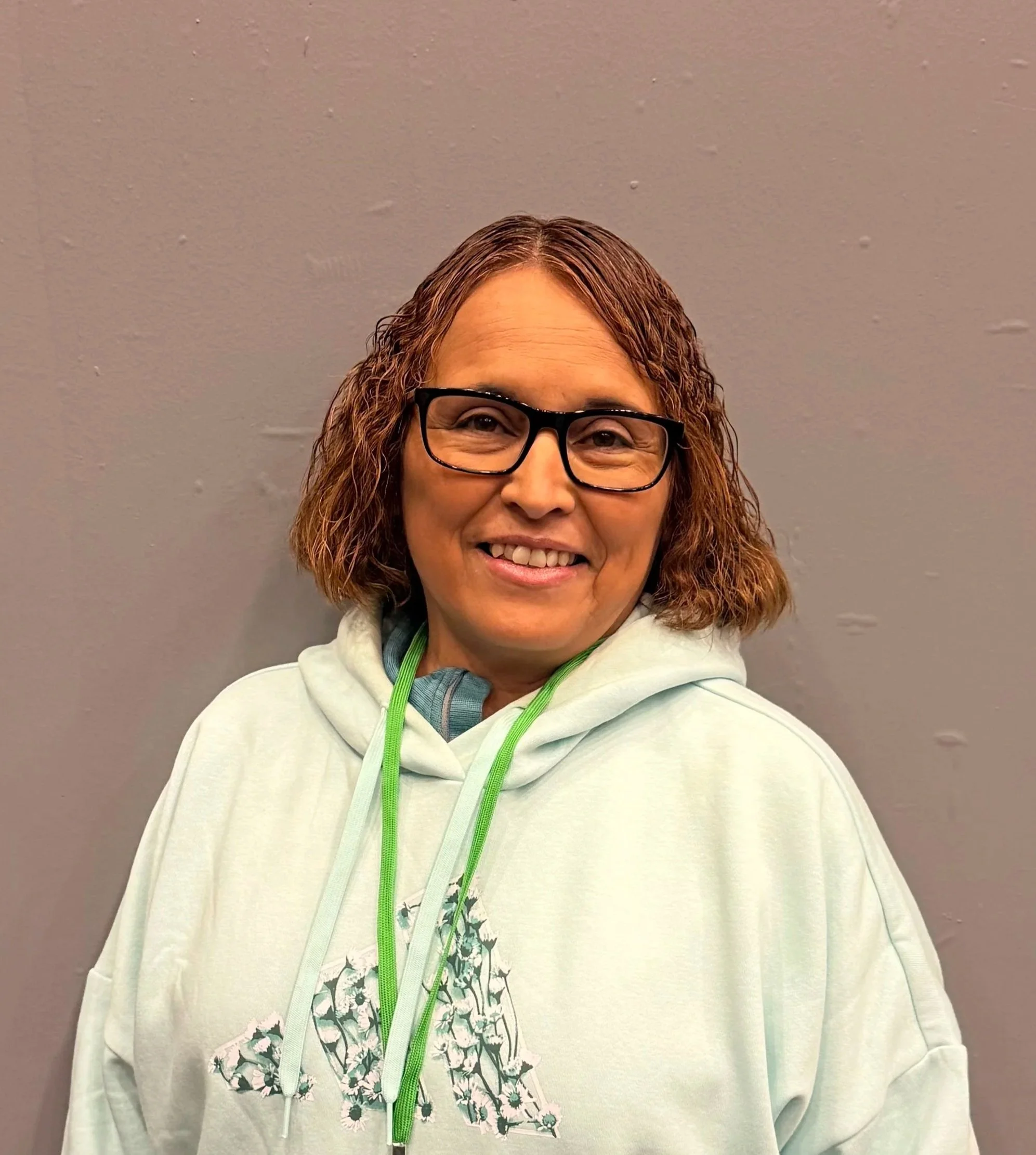A woman with curly brown hair, glasses, and a white hoodie smiling at the camera, standing against a gray wall.
