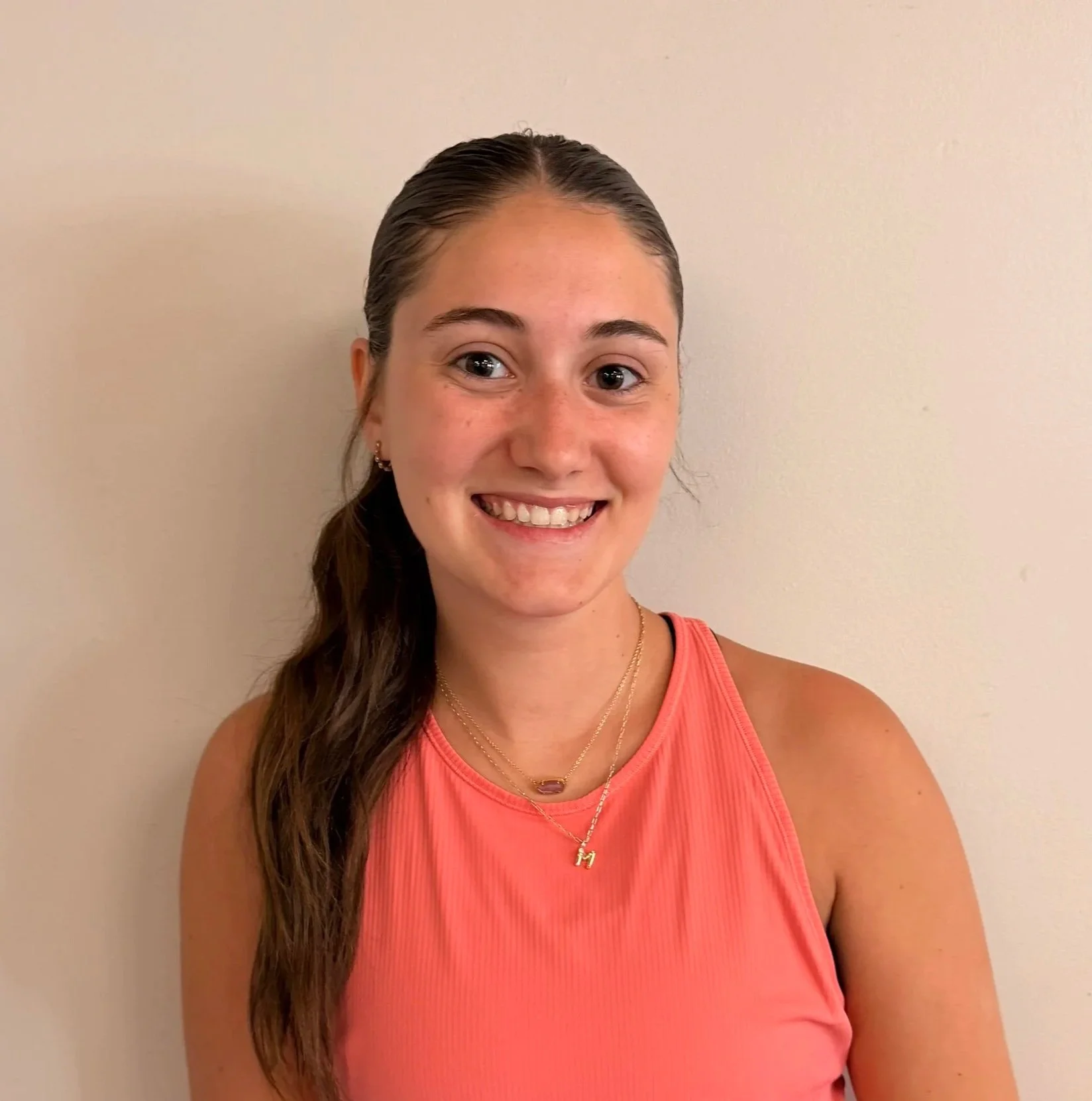 Smiling woman with long wavy hair in a ponytail, wearing a pink sleeveless top, gold necklaces, earrings, and a pendant with the letter "M", standing against a plain beige wall.