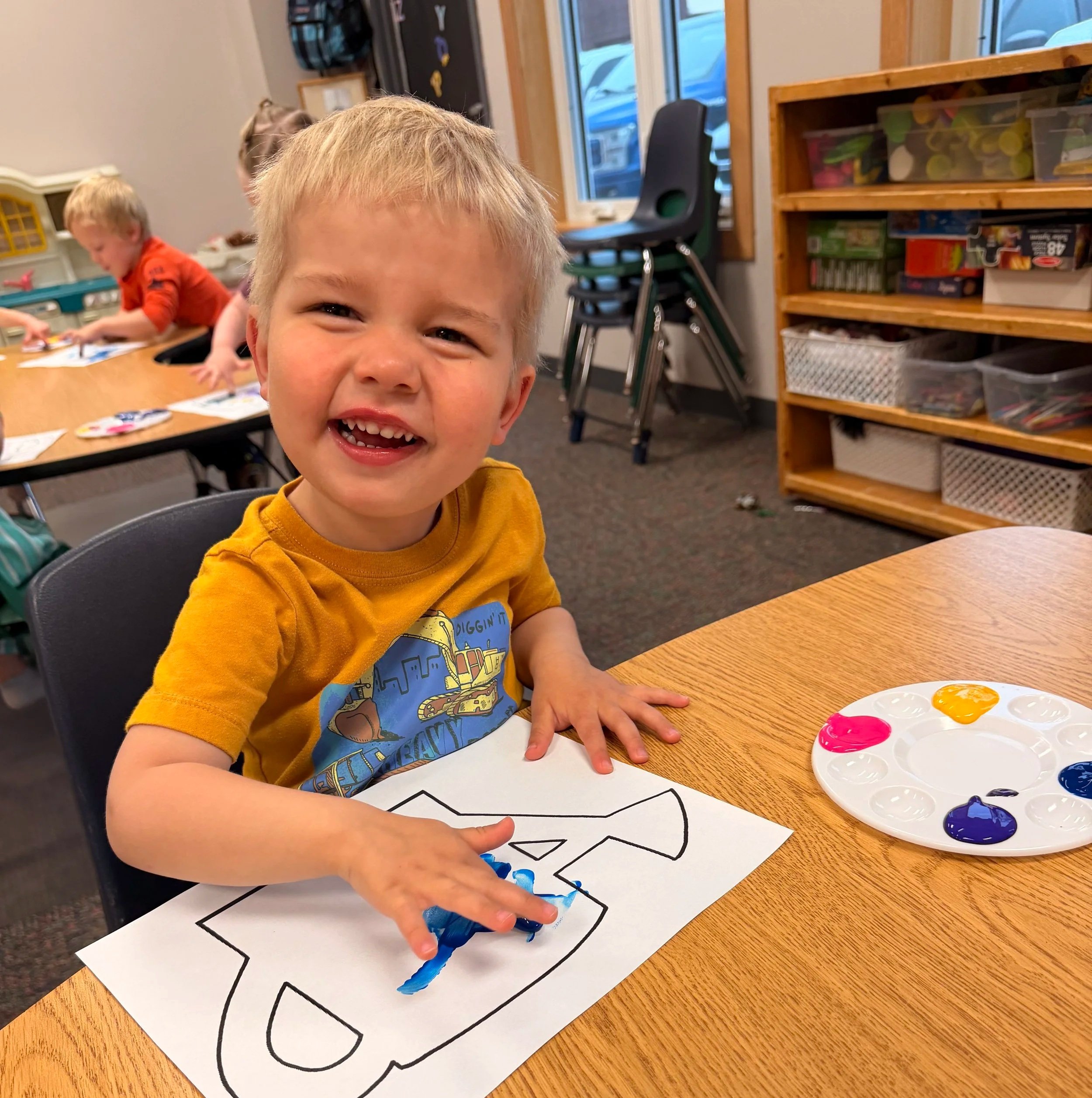 A young boy with blond hair smiling and pointing to a large coloring sheet on the table, with children in the background working on similar activities, in a classroom setting.