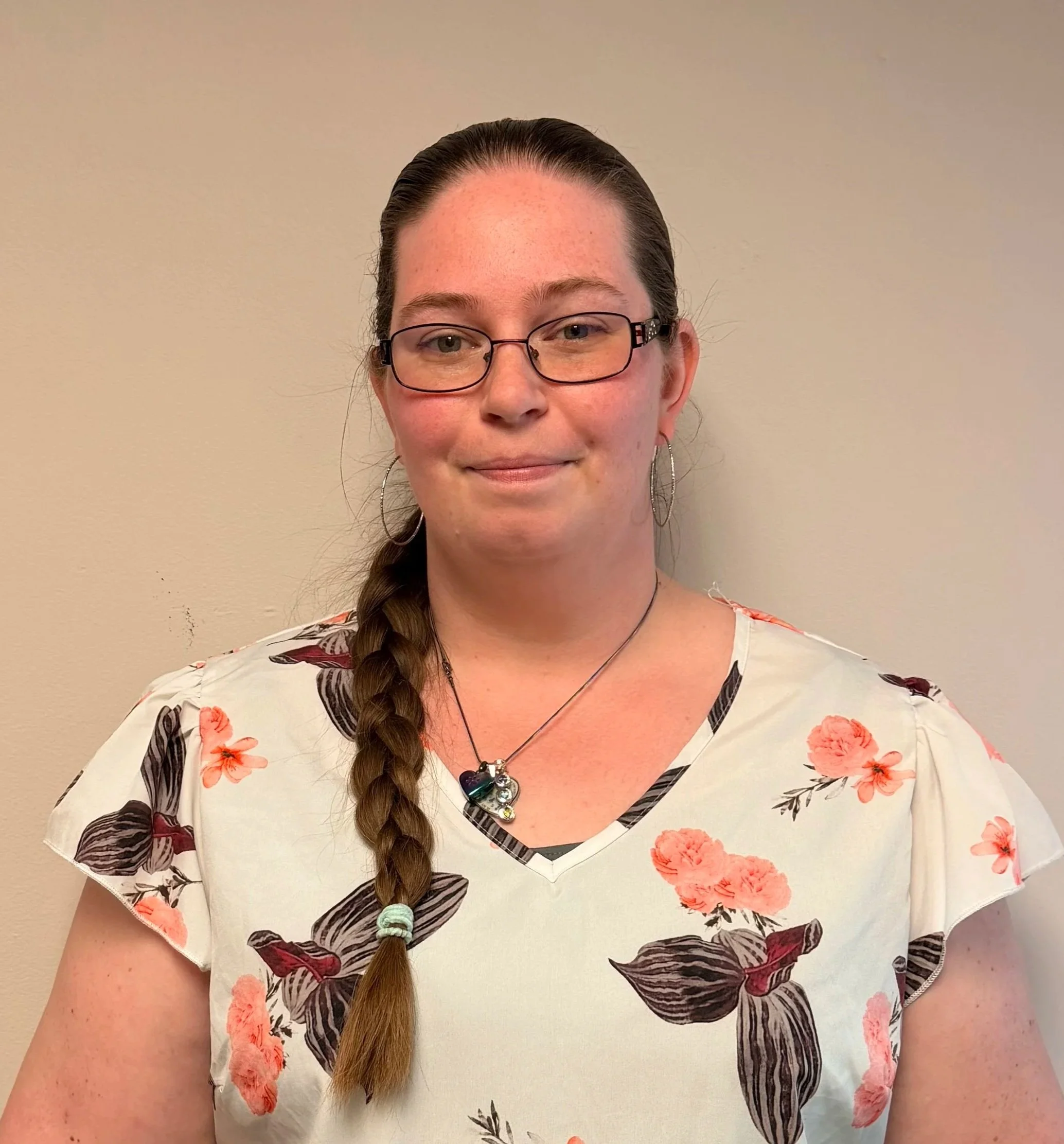A woman with glasses and long brown hair in a braid stands in front of a beige wall. She's wearing a floral-patterned shirt with pink and red flowers and dark leaves, and a necklace with multiple pendants, including a shell and a stone. The lighting is soft and warm.