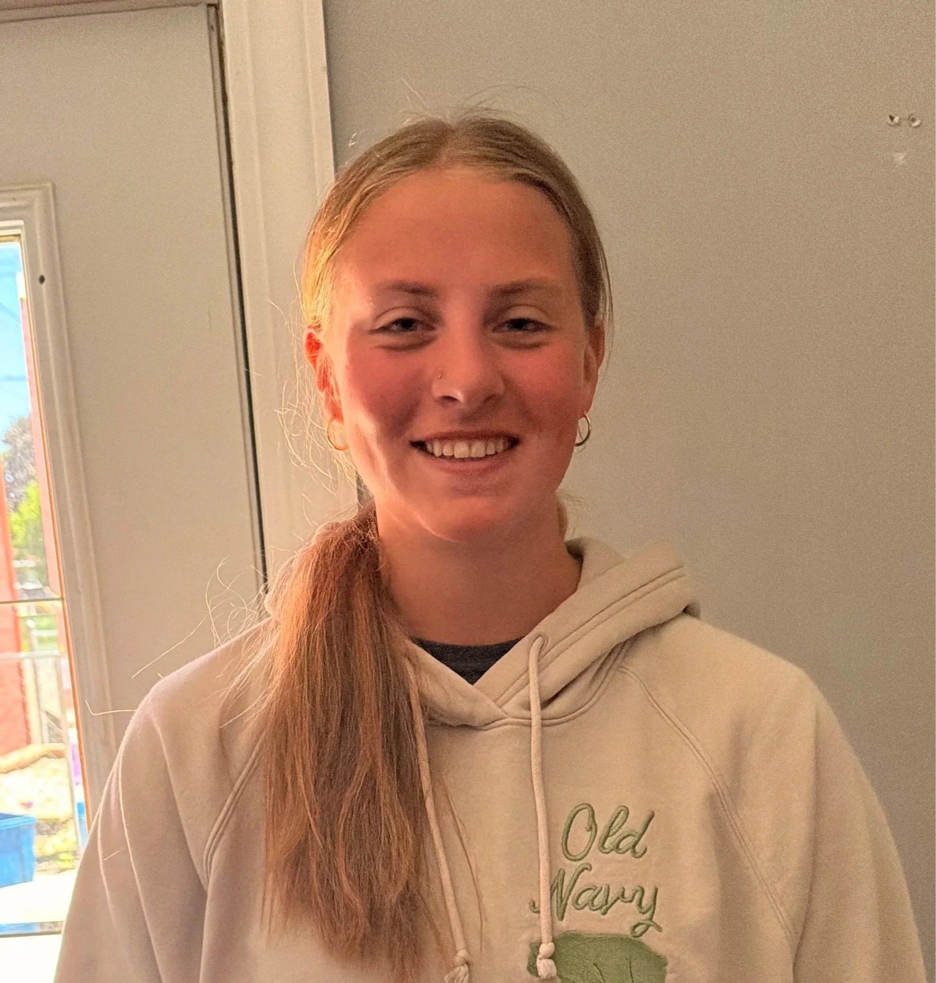 Close-up of a young woman with long red hair smiling, wearing a beige hoodie with green text and a leaf graphic, standing indoors near a window with a gray wall background.