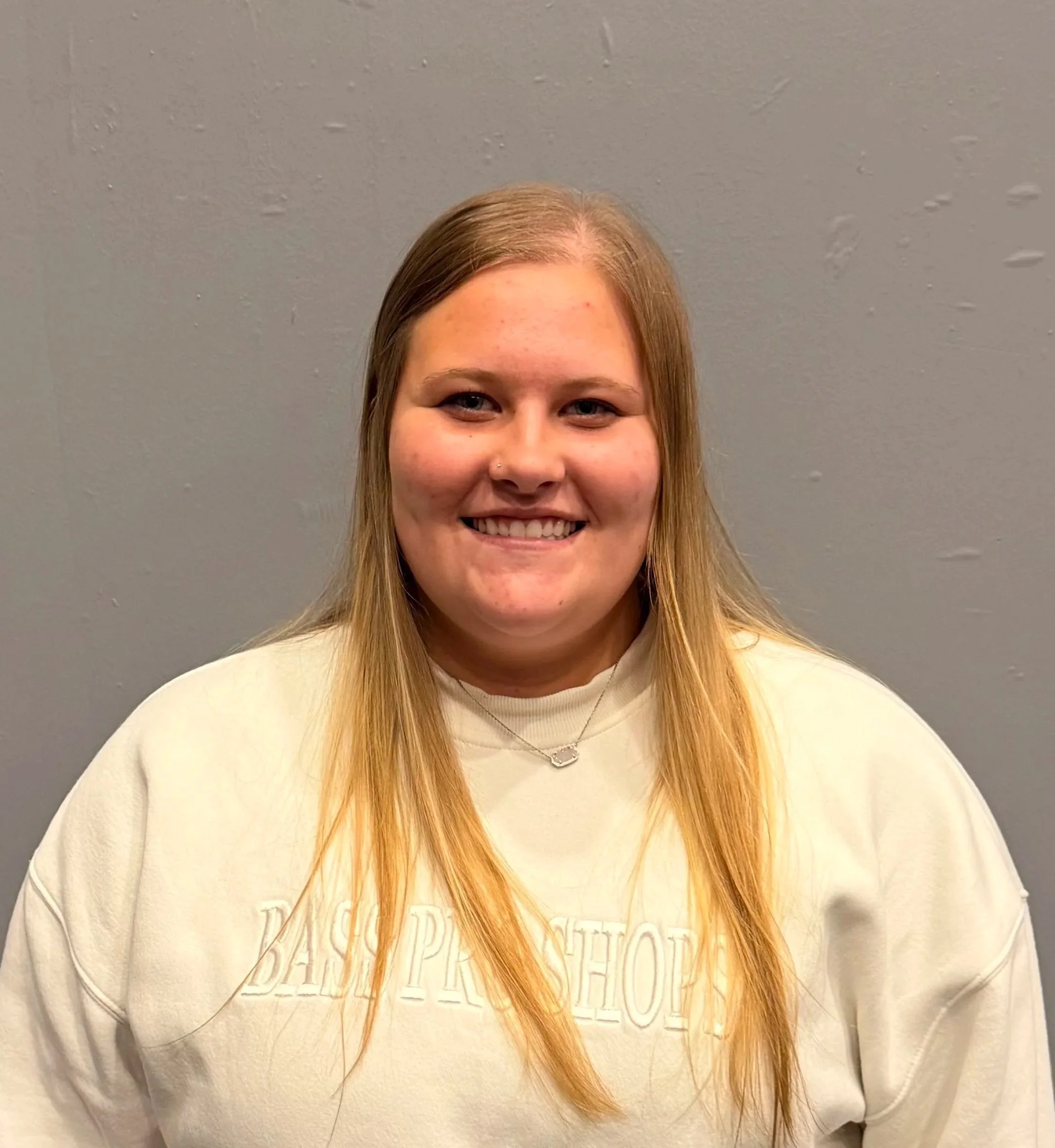 Close-up of a smiling woman with long blonde hair, wearing a cream-colored sweatshirt and a delicate silver necklace, standing against a gray textured wall.
