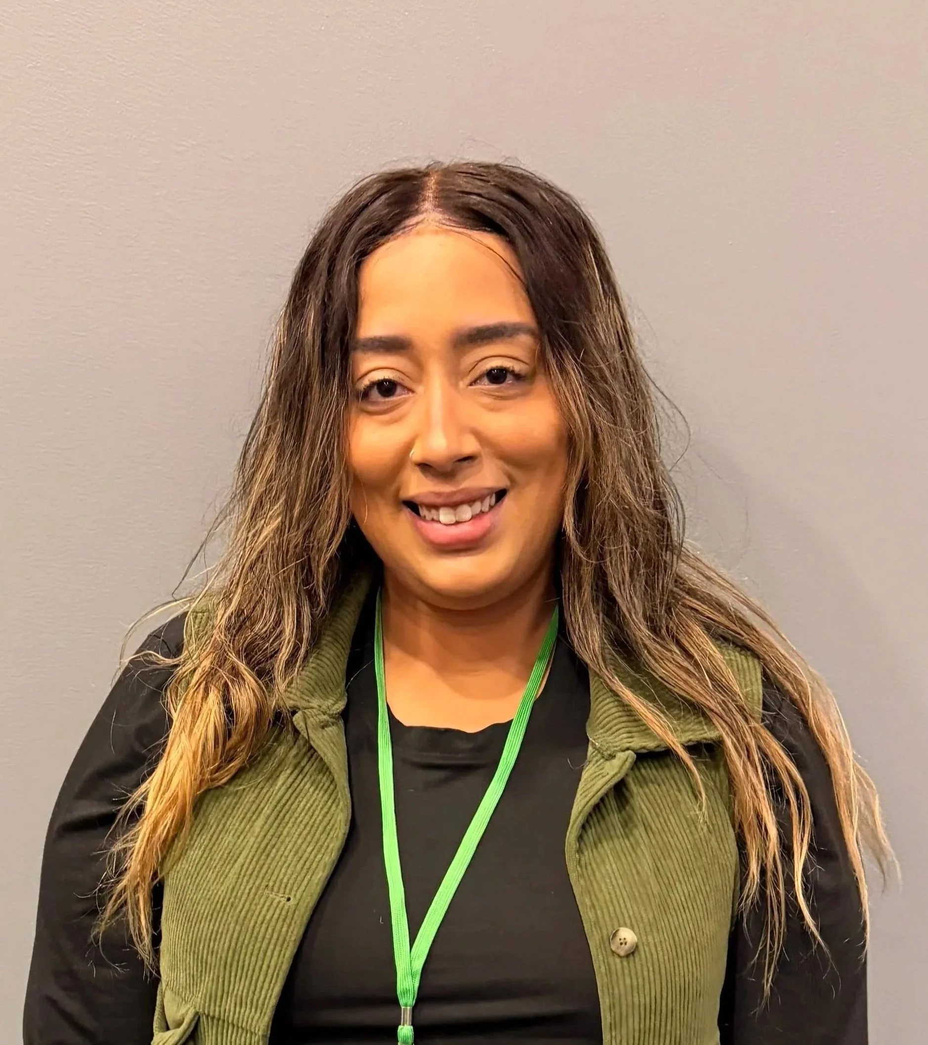 Close-up of a woman with long wavy brown hair, smiling, wearing a black shirt, a green vest, and a green lanyard against a plain gray background.