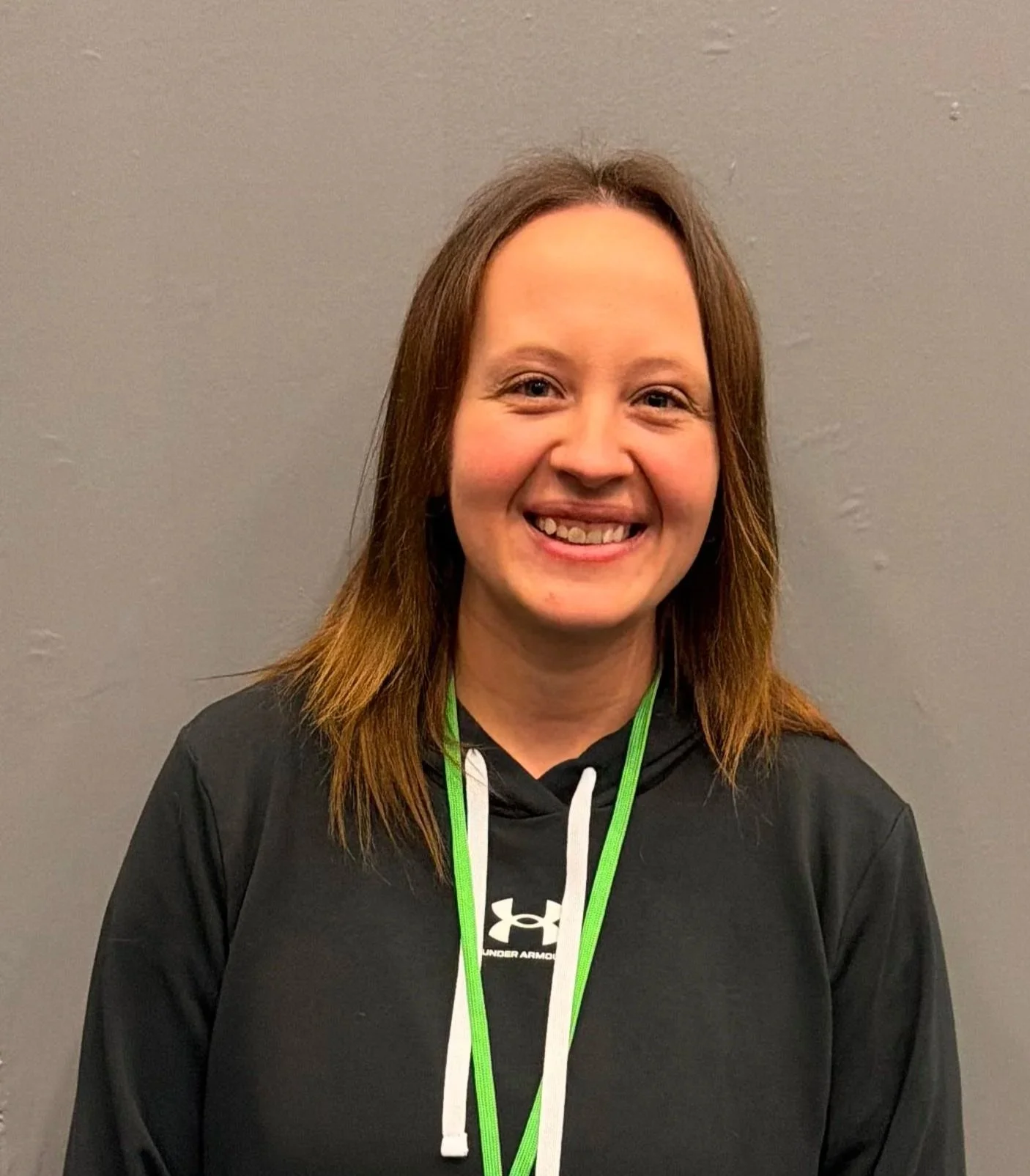 A woman with shoulder-length brown hair smiling, wearing a black Under Armour hoodie with white drawstrings, and a green lanyard around her neck, standing against a plain gray wall.