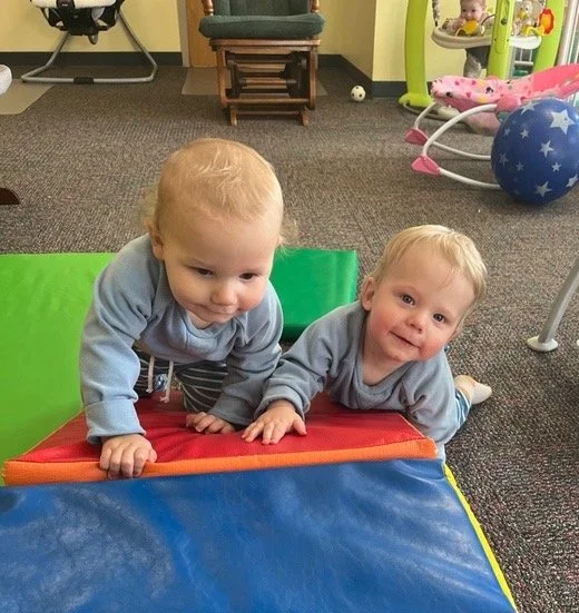 Two young boys crawling on a padded play mat indoors, with baby toys and furniture in the background.